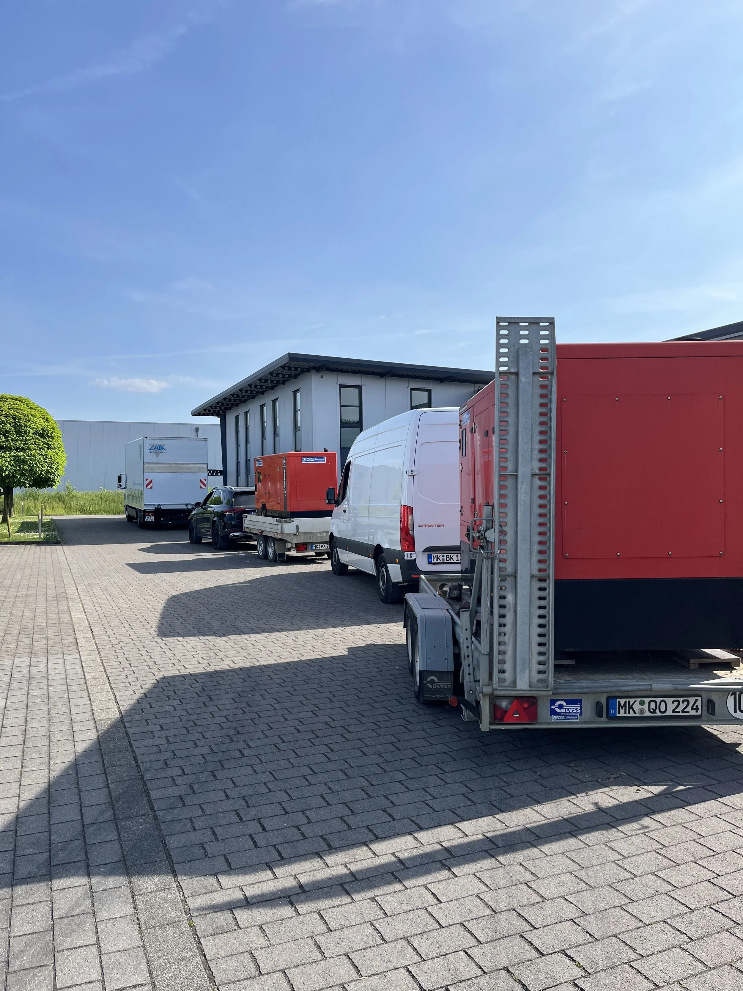 Several parked vehicles, including vans, trucks, and trailers, in a parking lot outside a modern building on a sunny day with a clear blue sky.
