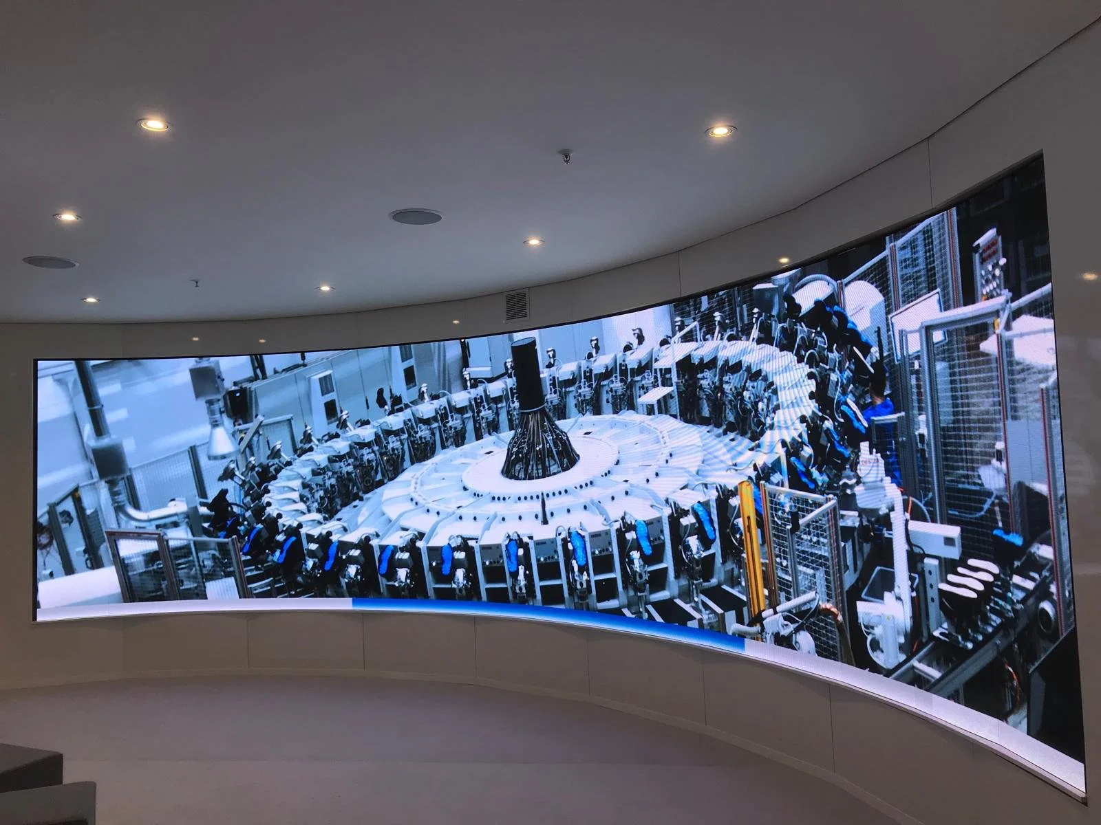 Curved digital display showing an industrial automation or robotic system with machinery and robotic arms in a high-tech factory setting.