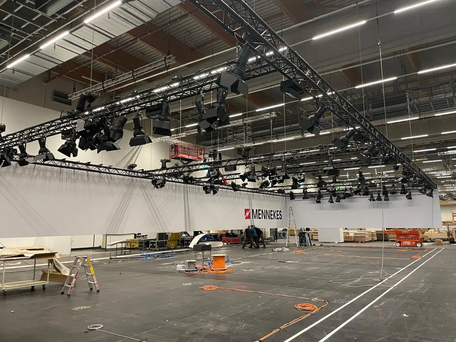 An indoor event space or gymnasium under setup, with a large white wall bearing the 'Mennekes' logo, ceiling-mounted lighting and equipment trusses, some workers and ladders, and construction materials and tools scattered on the floor.