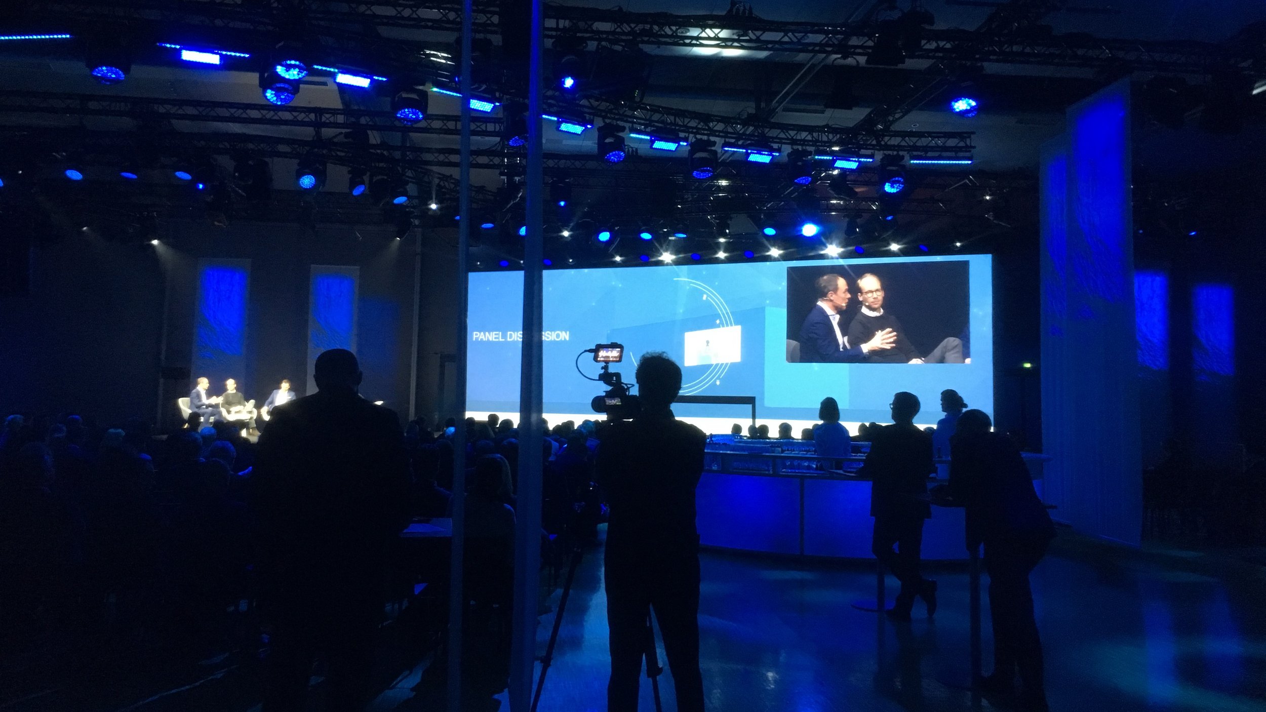 A stage with a large screen displaying a panel discussion, two women speaking on stage, and a dark room with audience members and camera operators.