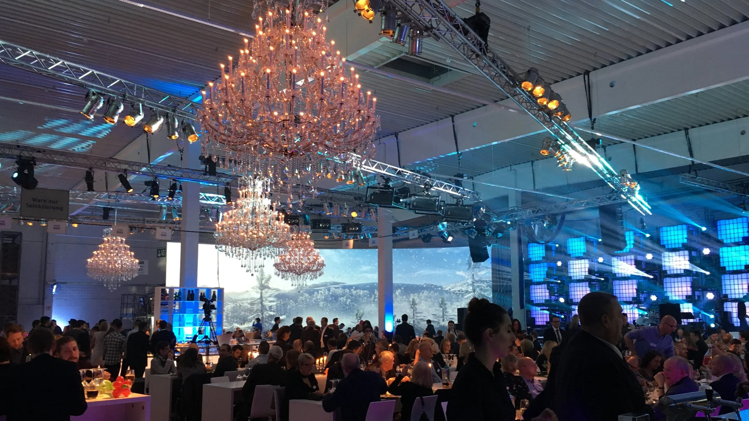 An indoor event with chandeliers hanging from the ceiling, large stage with blue lighting and screens, and many people seated and standing, enjoying entertainment and socializing.