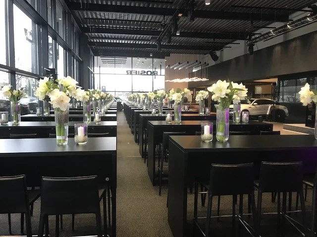 Interior of a modern, well-lit event space with black tables, each decorated with tall vases of white flowers and candles, set up for a formal gathering or reception.