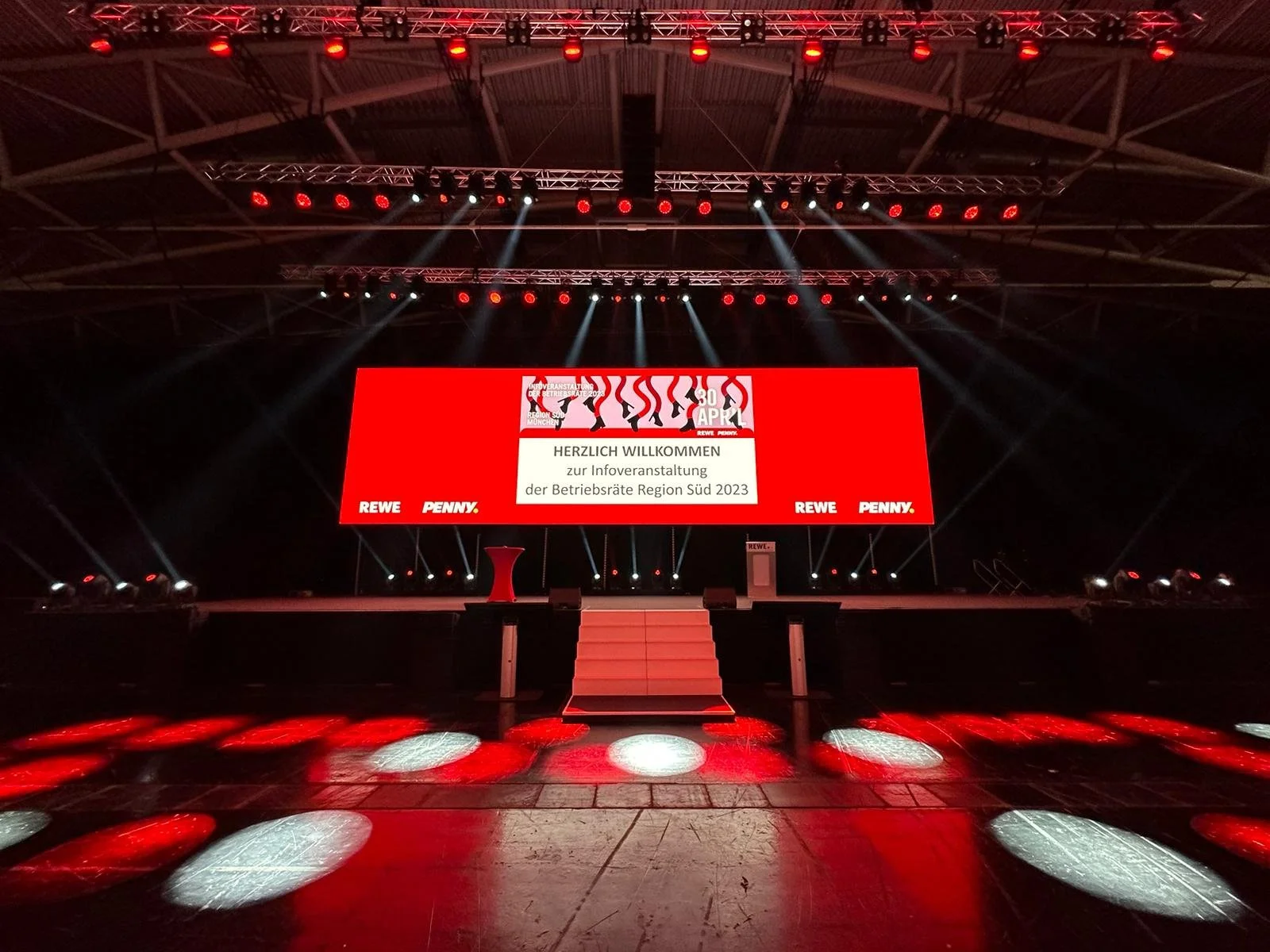 Empty stage with a large digital screen displaying welcome message in German, two small podiums, and stage lighting, set up for an event in a large indoor venue.