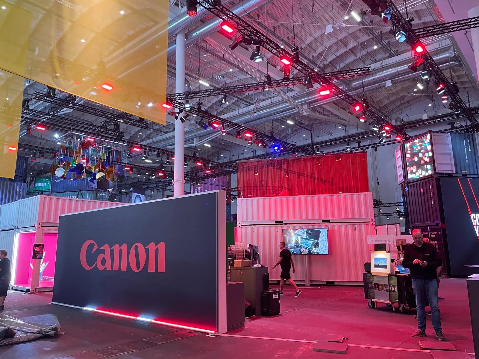 Interior of a trade show or convention center with various booths, including a large Canon display with pink and black branding, hanging colorful art pieces, and fluorescent lighting.