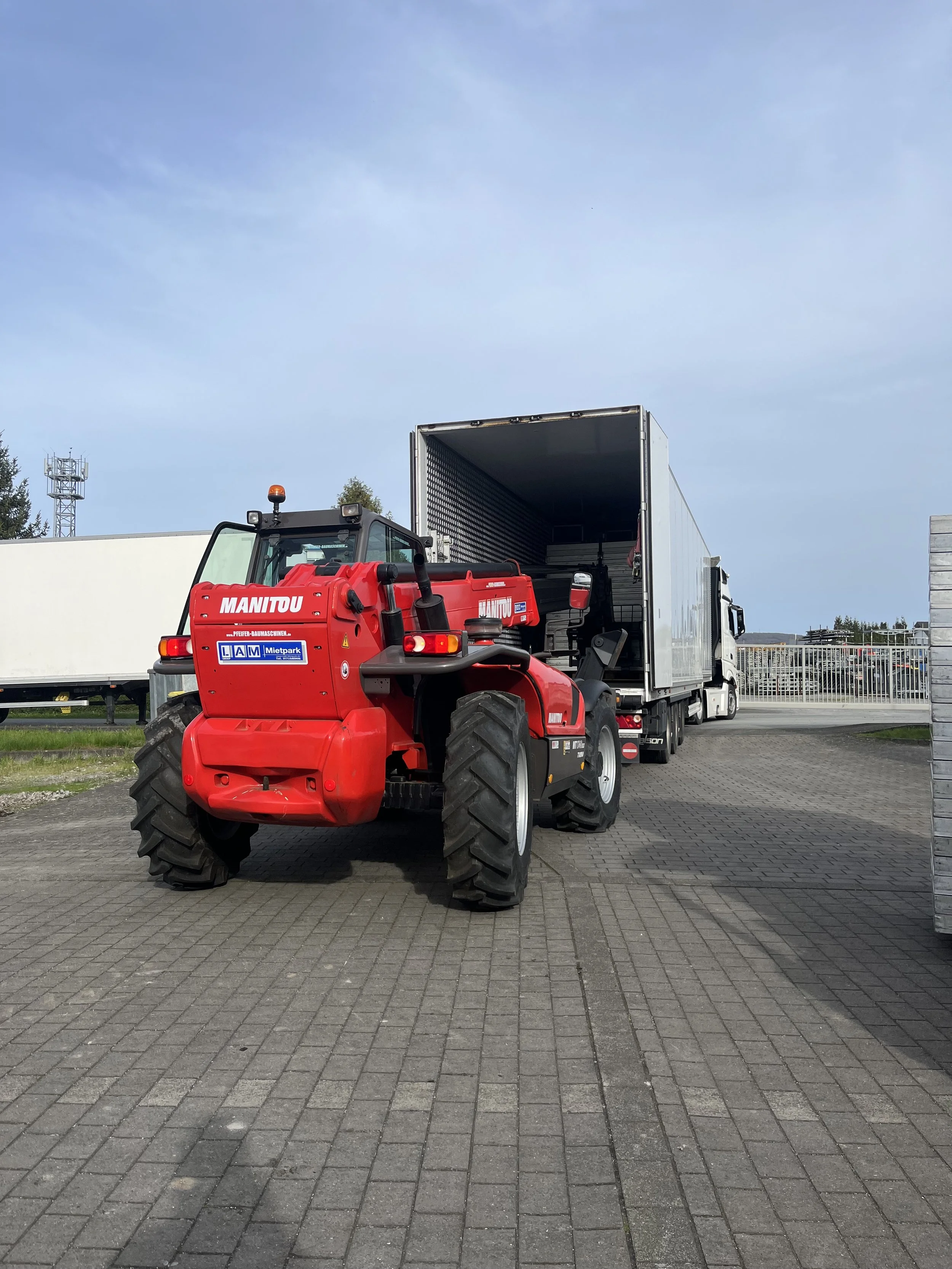 A red Manitou telehandler vehicle is being loaded onto a white truck with an open cargo area in an outdoor industrial area. The ground is paved with interlocking bricks and there are some trees and a fence in the background.