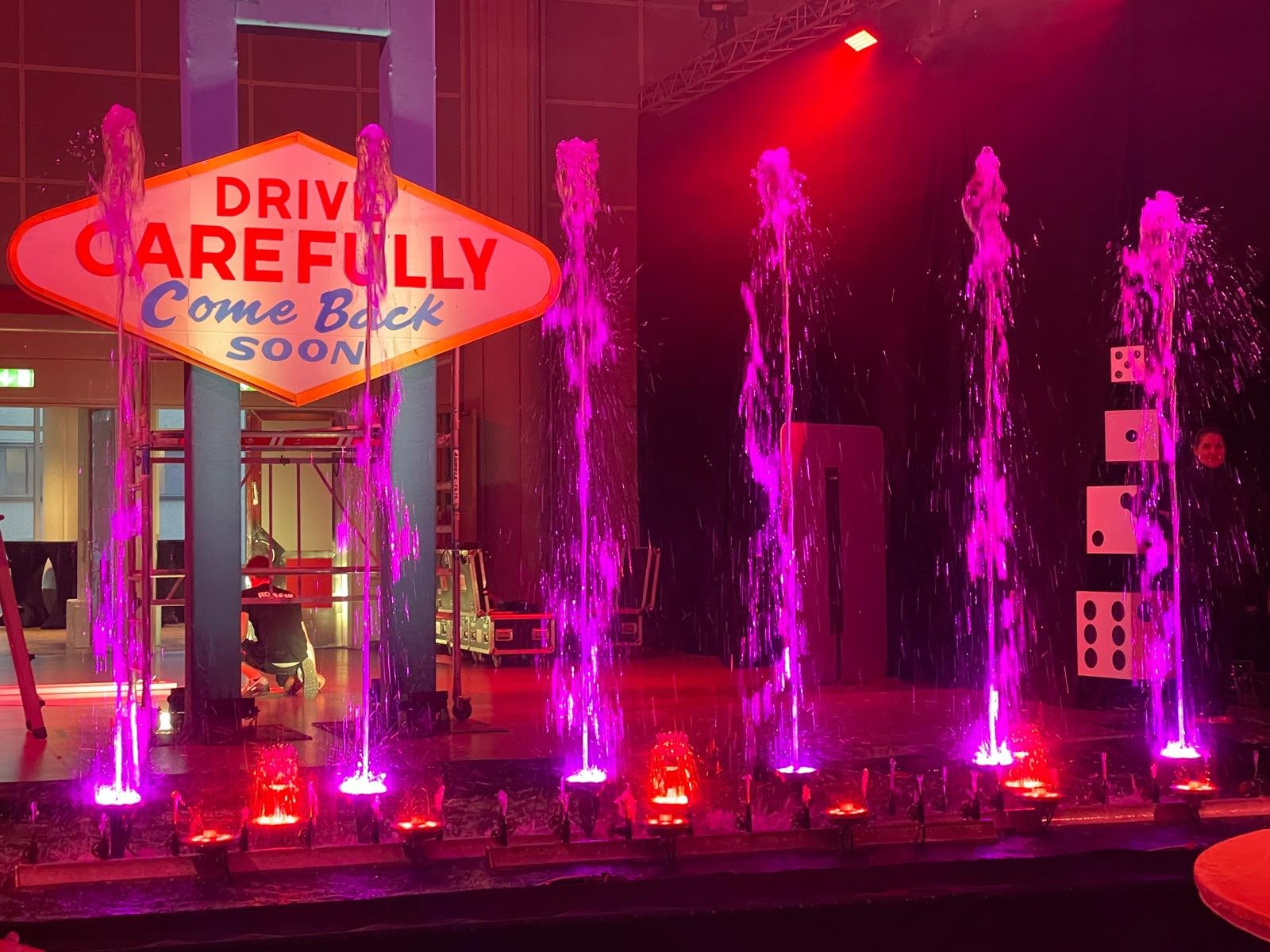 Indoor water fountain with pink lighting and a sign that reads 'Drive Carefully Come Back Soon' in an indoor space with a high ceiling.