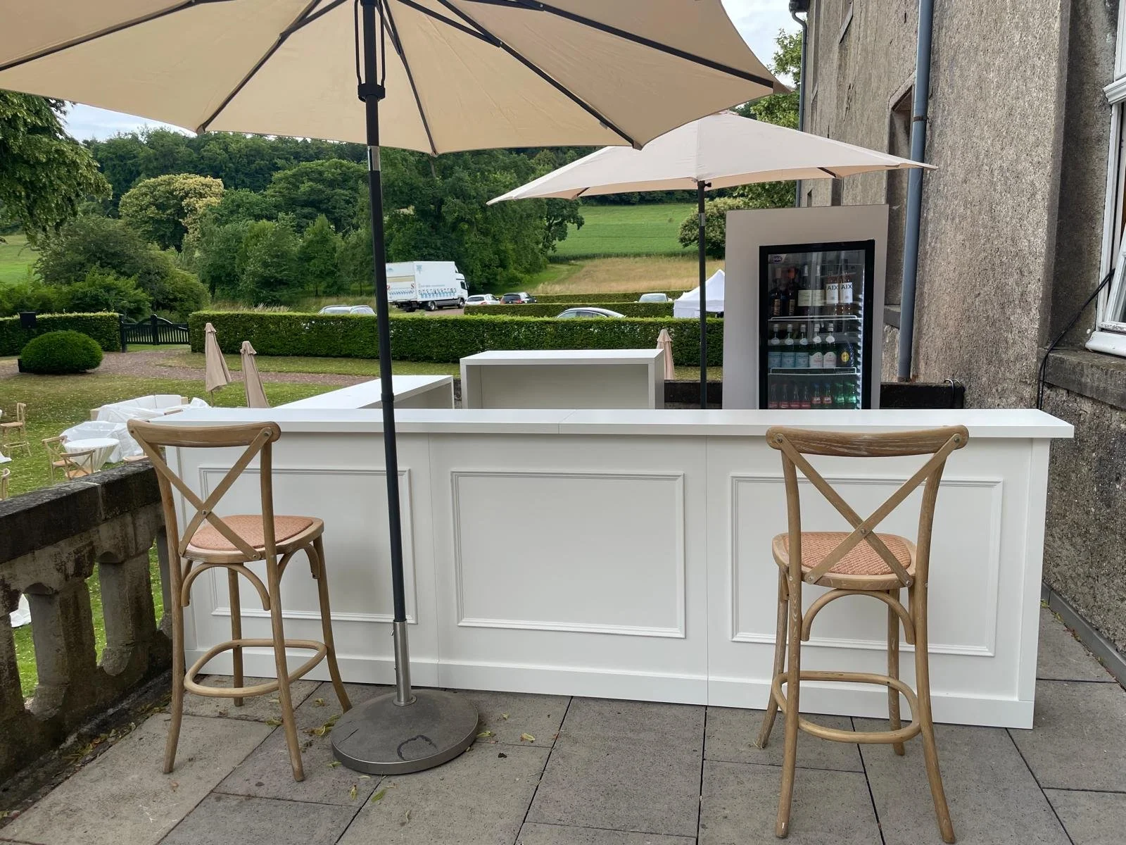 An outdoor bar setup with a white bar counter, two wooden bar stools with beige seats, two umbrellas providing shade, and a glass-front refrigerator stocked with beverages, against a background of green fields and trees.
