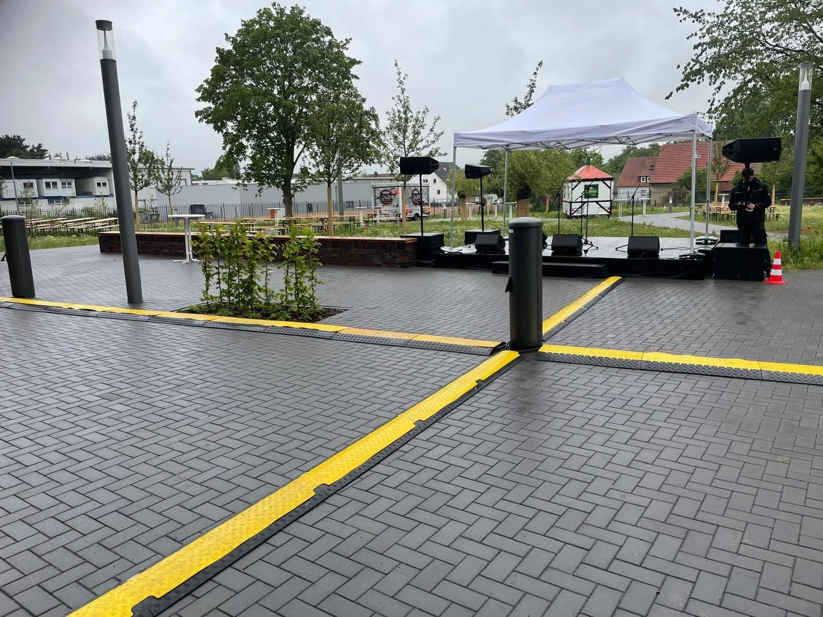 Empty outdoor stage with microphones, speakers, and a white canopy in a park, with a paved area, small plants, trees, and residential buildings in the background.