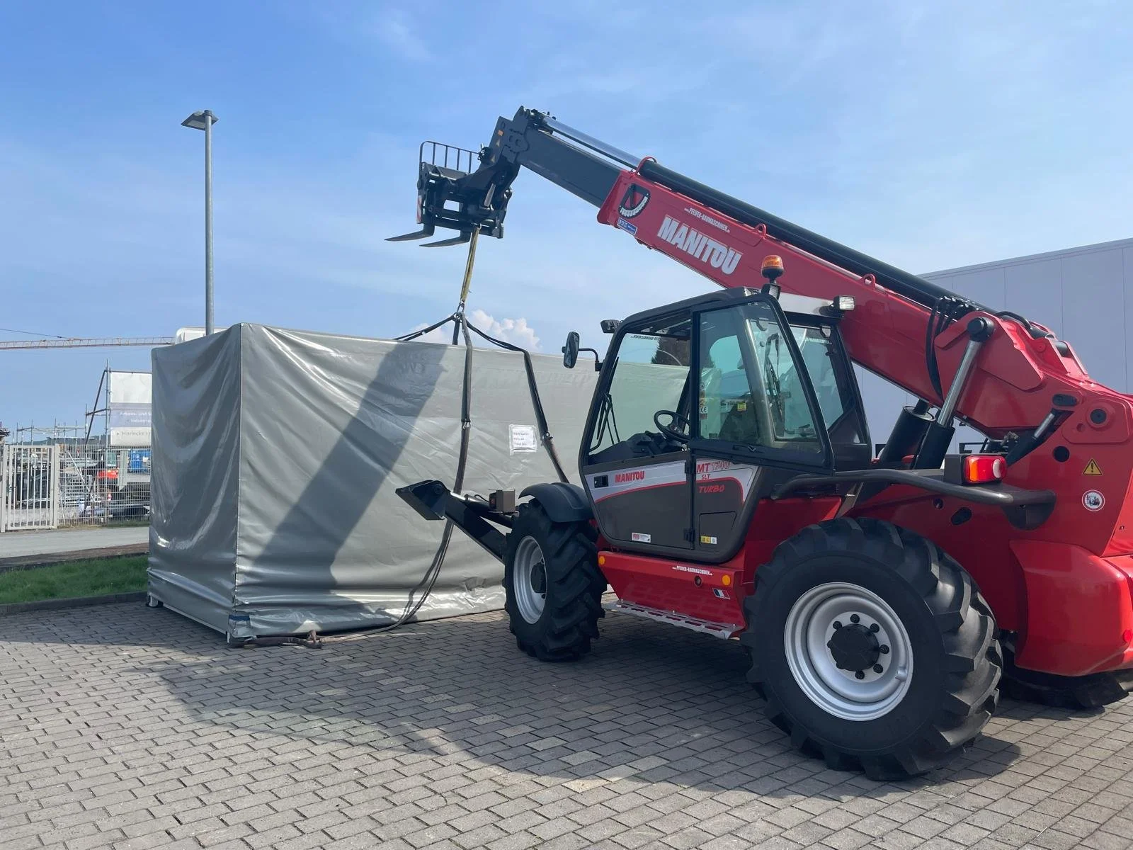 A red Manitou telescopic handler lifting a large covered object in an outdoor area with a paved surface, clear sky, and some facility structures in the background.