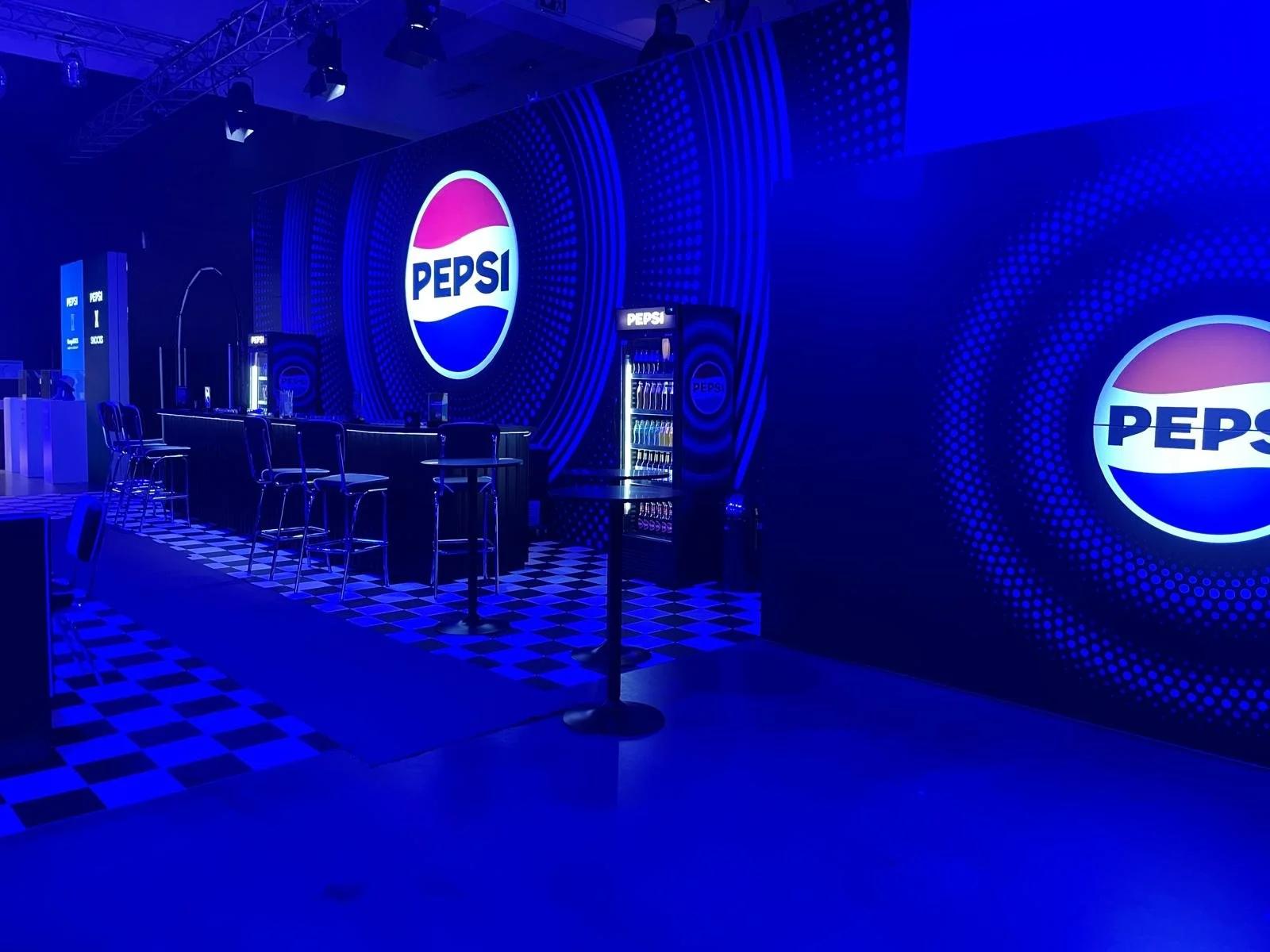A Pepsi-themed event space with multiple Pepsi logos on large screens, a bar with high chairs, and checkered flooring, illuminated by blue lighting.