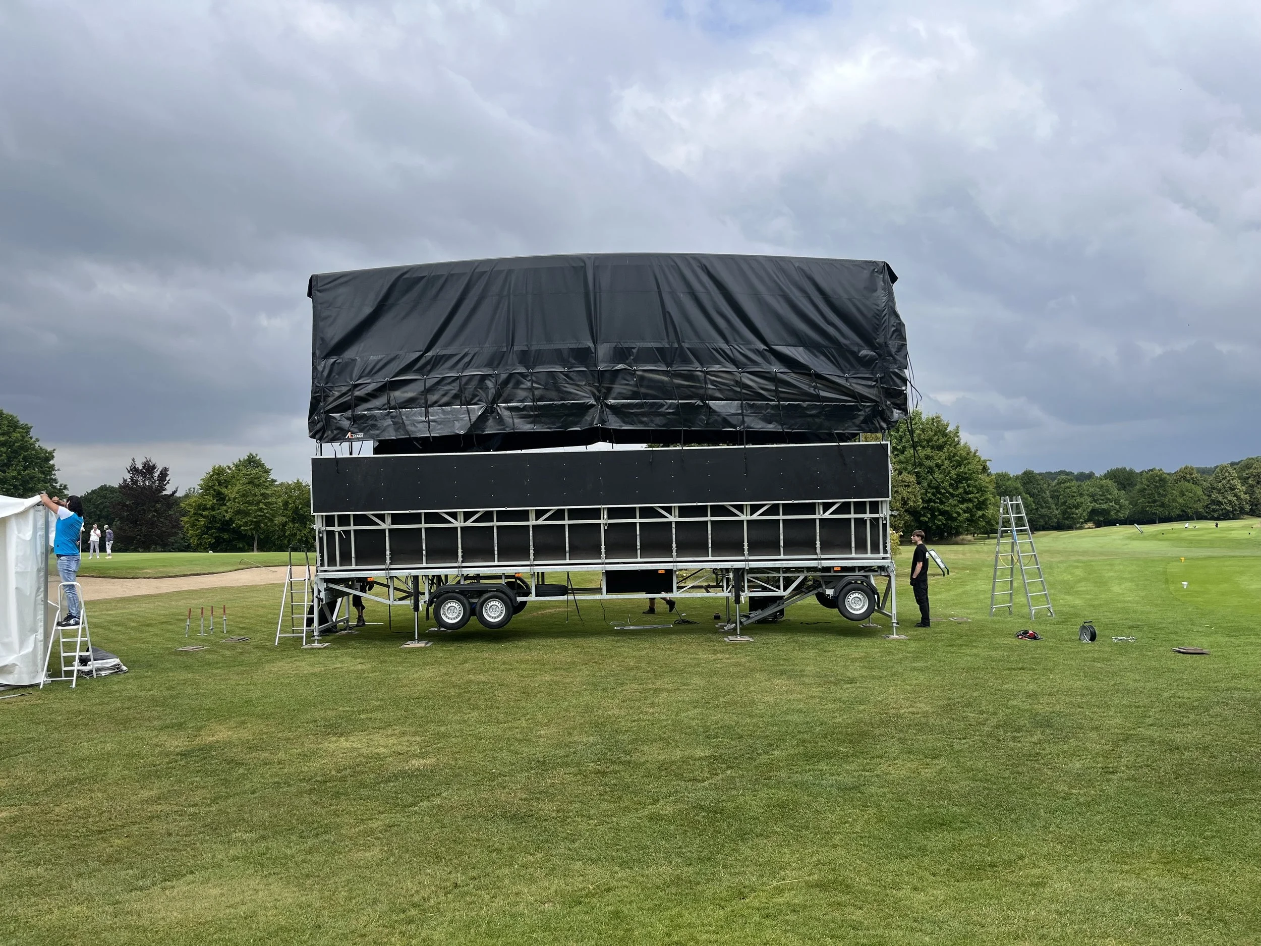 A large film or projection screen mounted on a mobile trailer on a golf course field, with people setting up equipment around it under cloudy skies.