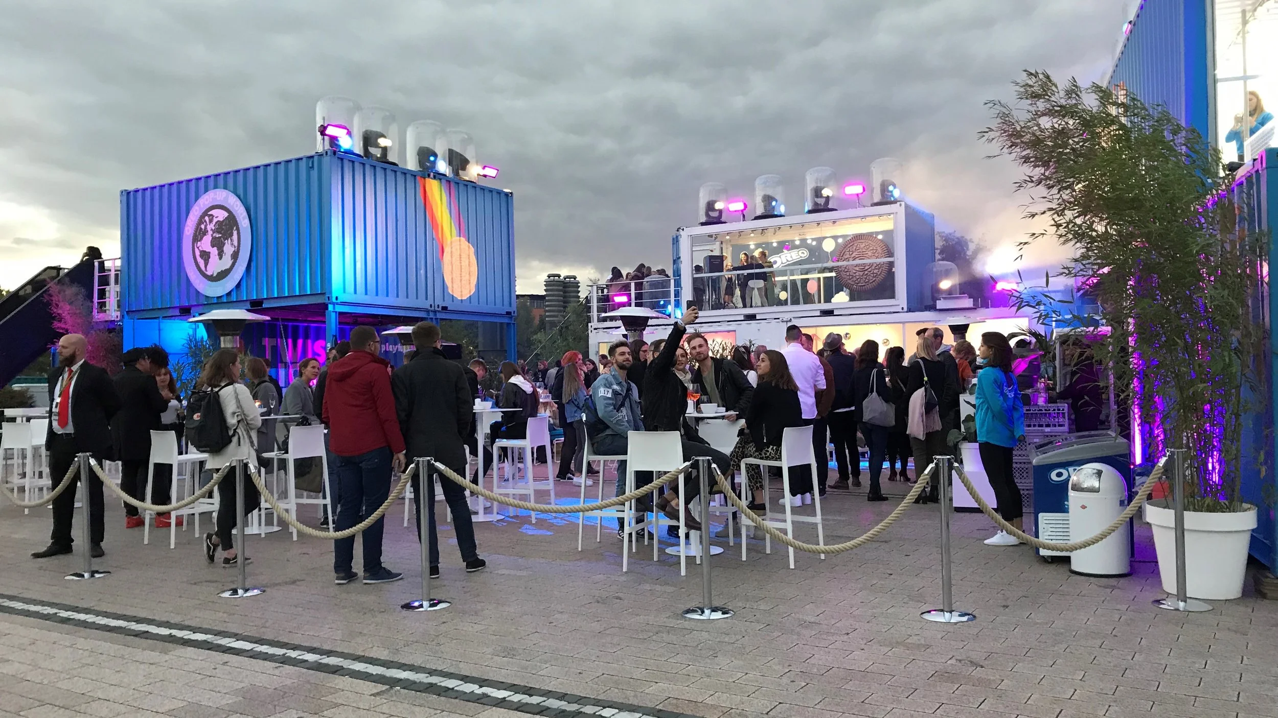 Outdoor event with people gathered around two colorful, illuminated shipping containers serving as bar and stage, with neon lighting, bar tables, and a cloudy sky overhead.
