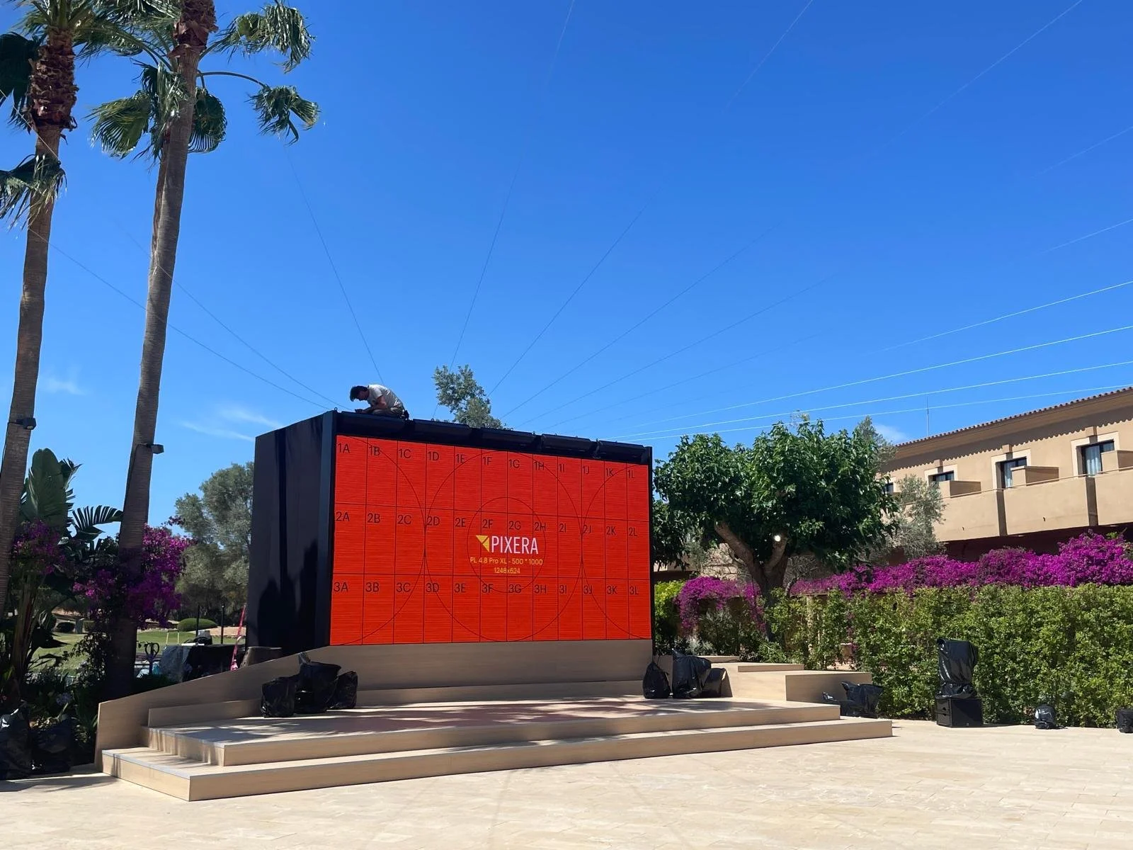 Outdoor stage with a large LED screen displaying the Pixera logo, surrounded by trees and flowering bushes, with a building in the background and a person working on top of the stage under a clear blue sky.