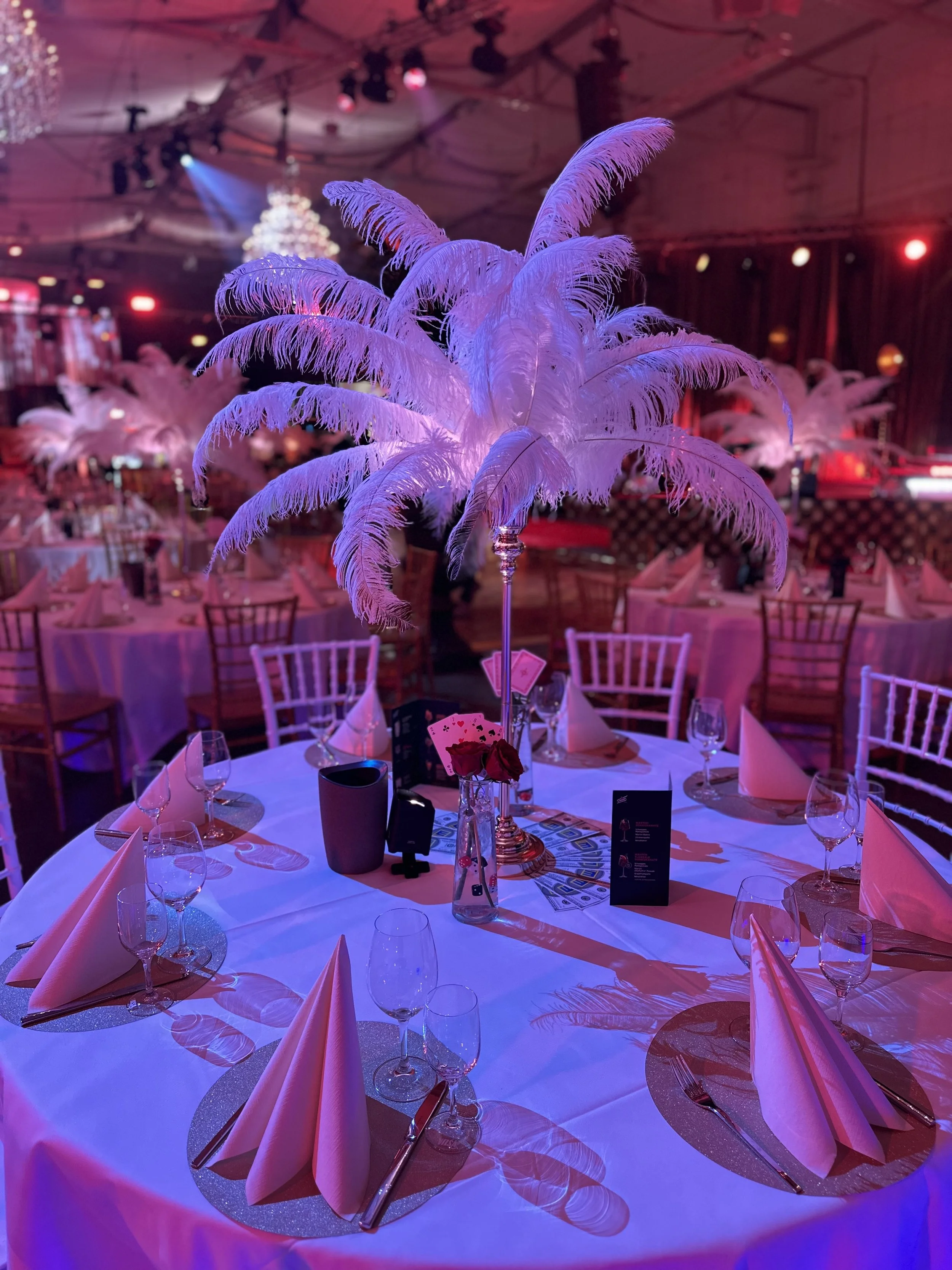 Elegant banquet table with white napkins, glassware, and a tall pink feather centerpiece, set in a dimly lit event hall with similar tables and purple and pink lighting.