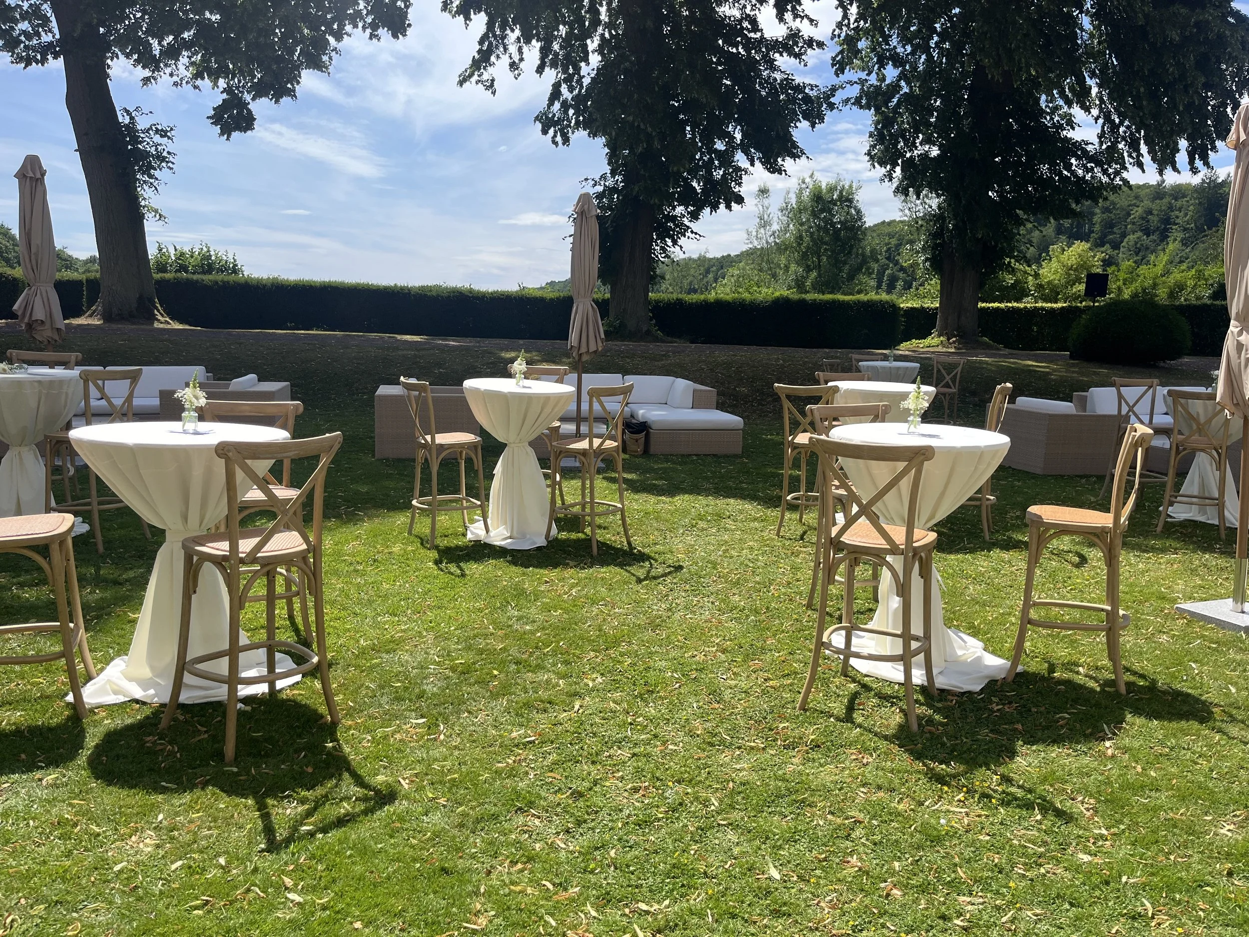 Outdoor garden event setup with round tables covered in white tablecloths, wooden chairs, parasols, and lounge seating under trees with a blue sky.