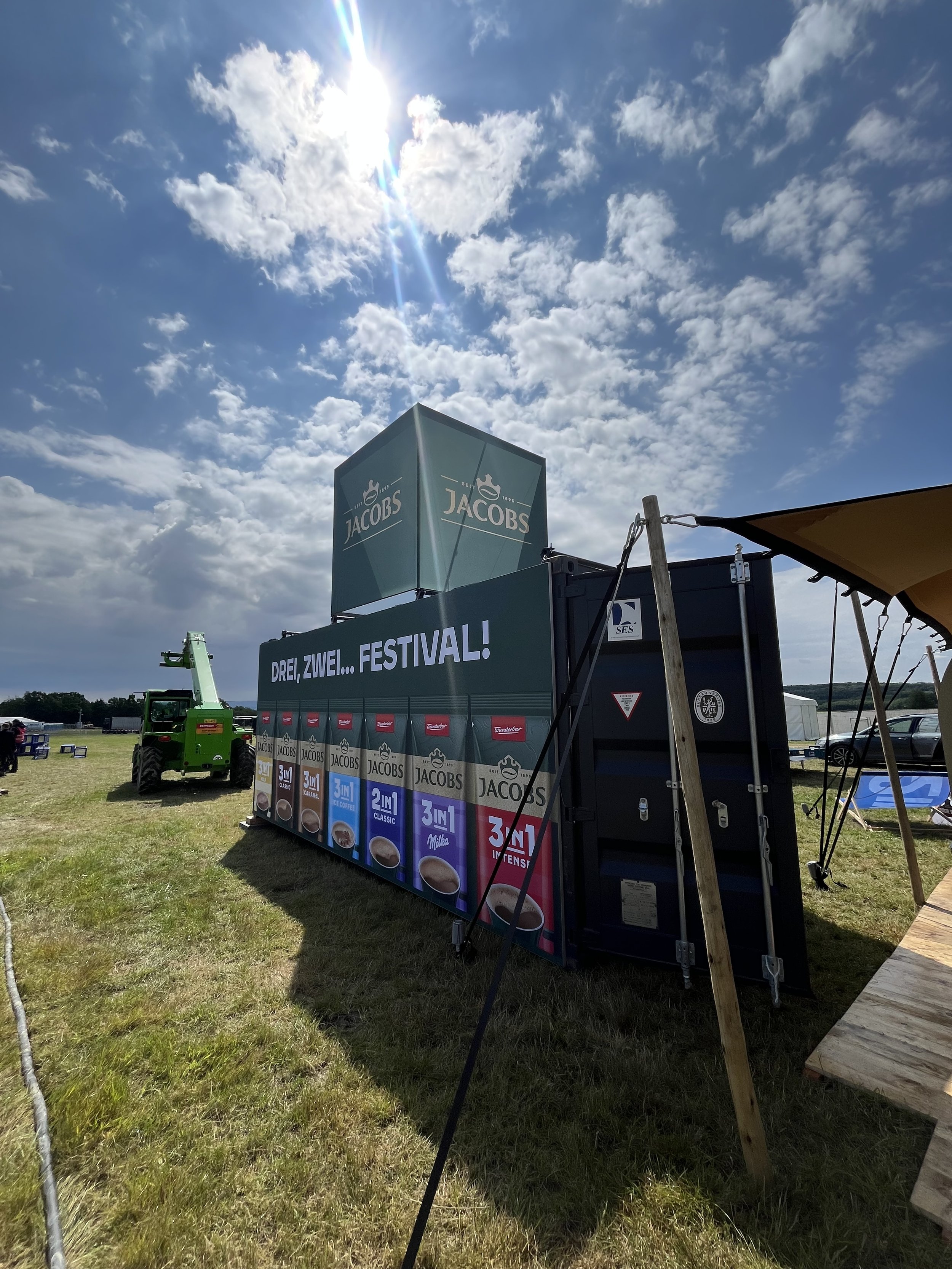 Outdoor festival scene with a large promotional container for Jacobs coffee, a green construction lift, and tents visible in the background, under a partly cloudy sky with sunlight.