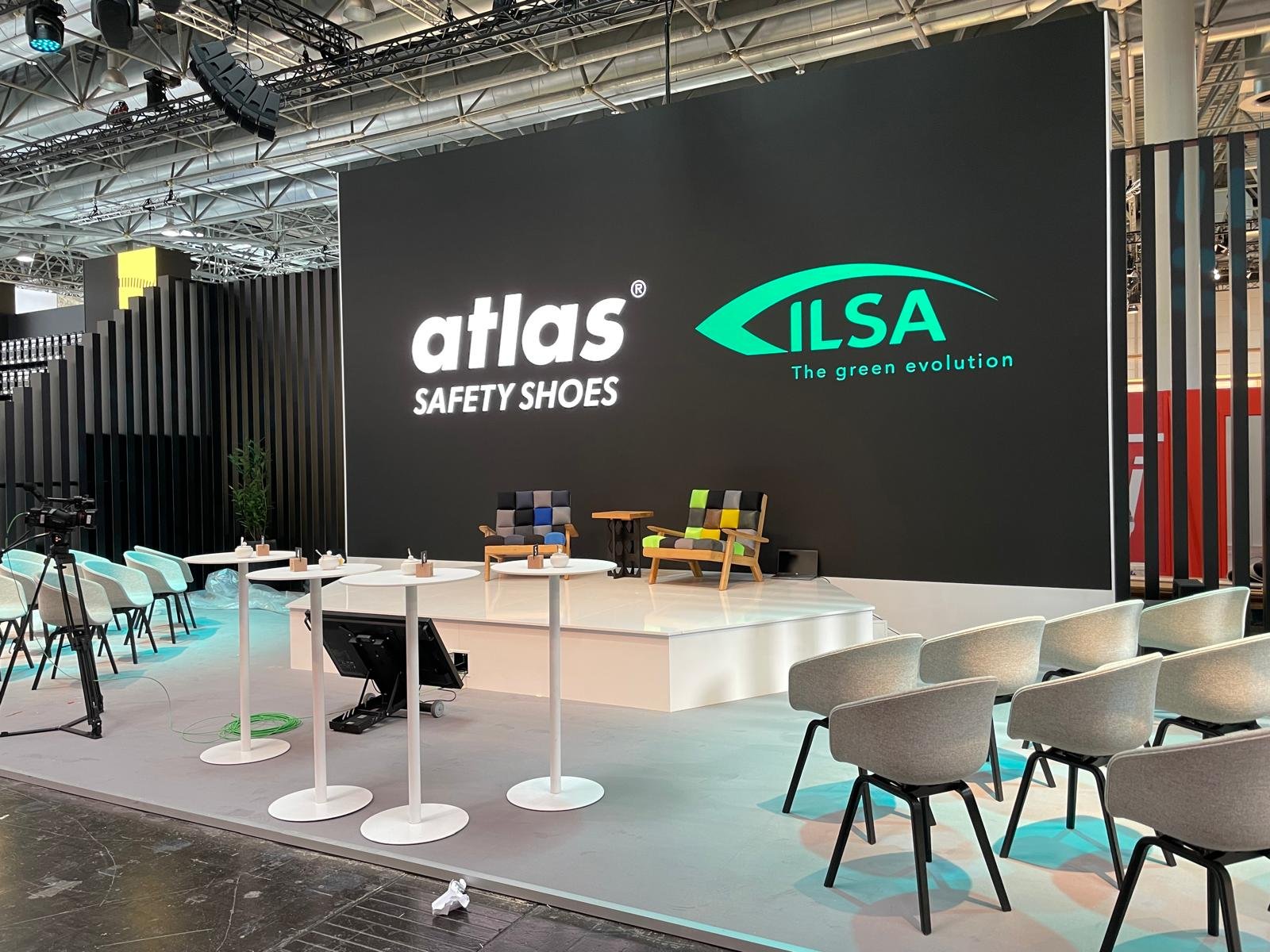 Stage setup at a trade show with a large black screen displaying the logos of Atlas Safety Shoes and ILSA The Green Evolution, surrounded by tables, chairs, and seating area.