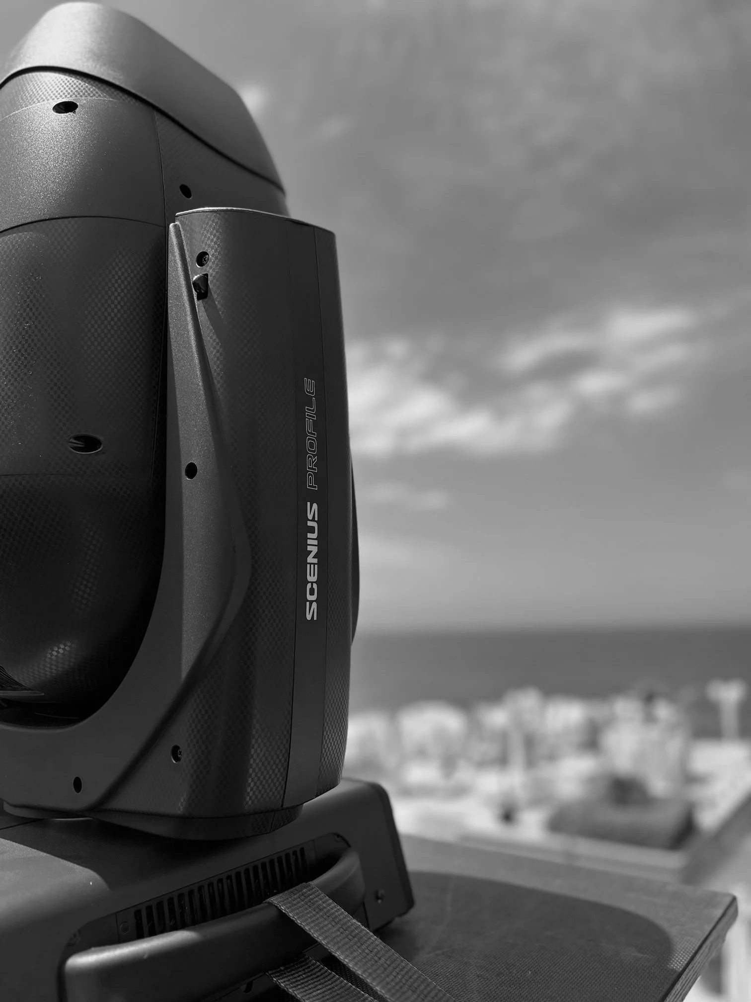 Close-up of a black professional speaker labeled 'SCENIUS PROFILE' on a rooftop with a city skyline and cloudy sky in the background.