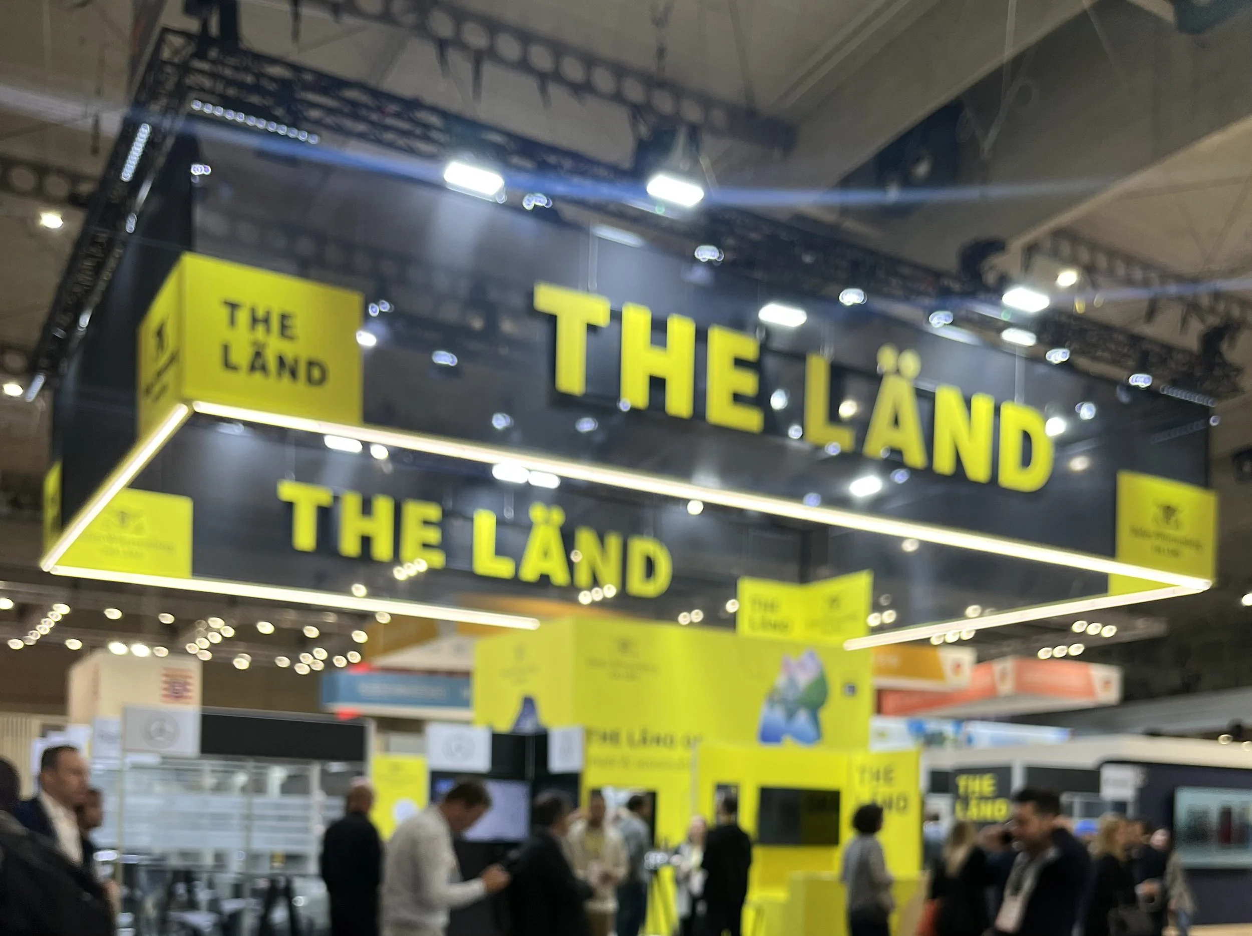 Exhibition booth with yellow signs that read "The Land" in bold black letters, with people walking and interacting around the booth.