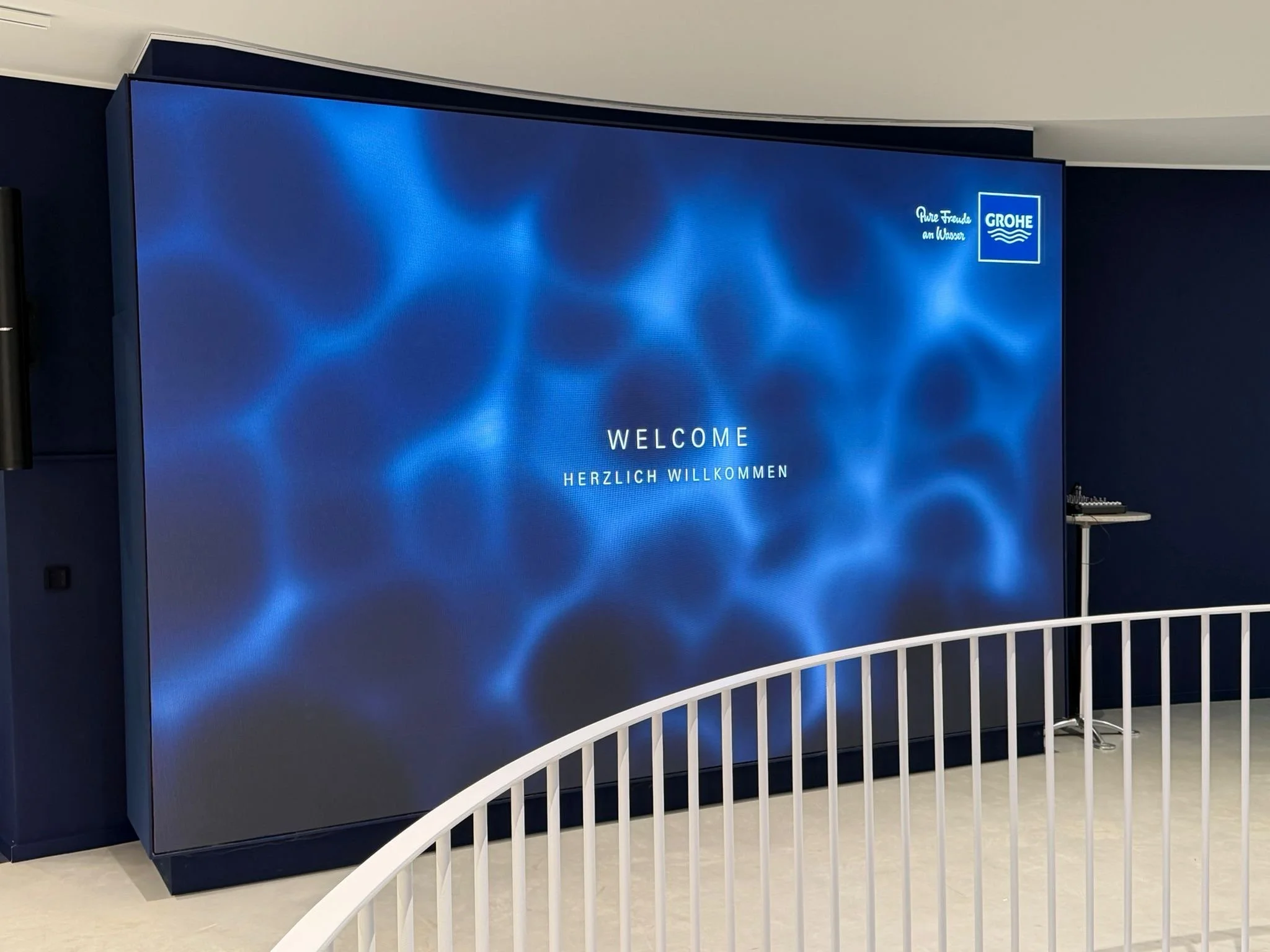 Large digital screen displaying a blue water ripple background with the text "Welcome" and "Herzlich Willkommen". The top right corner has the Grohe logo and the phrase "Pure Freude am Wasser". A white railing is in the foreground, and a small table 