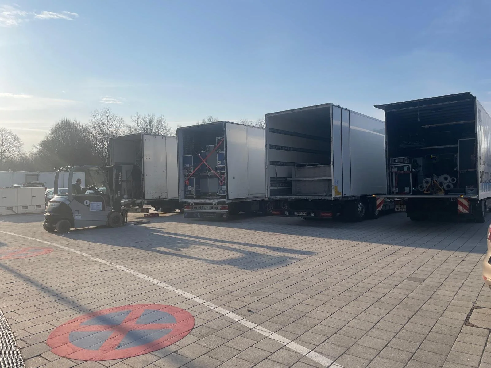 A small electric forklift moving a large freight container in a parking lot with other empty freight containers in the background and clear sky overhead.
