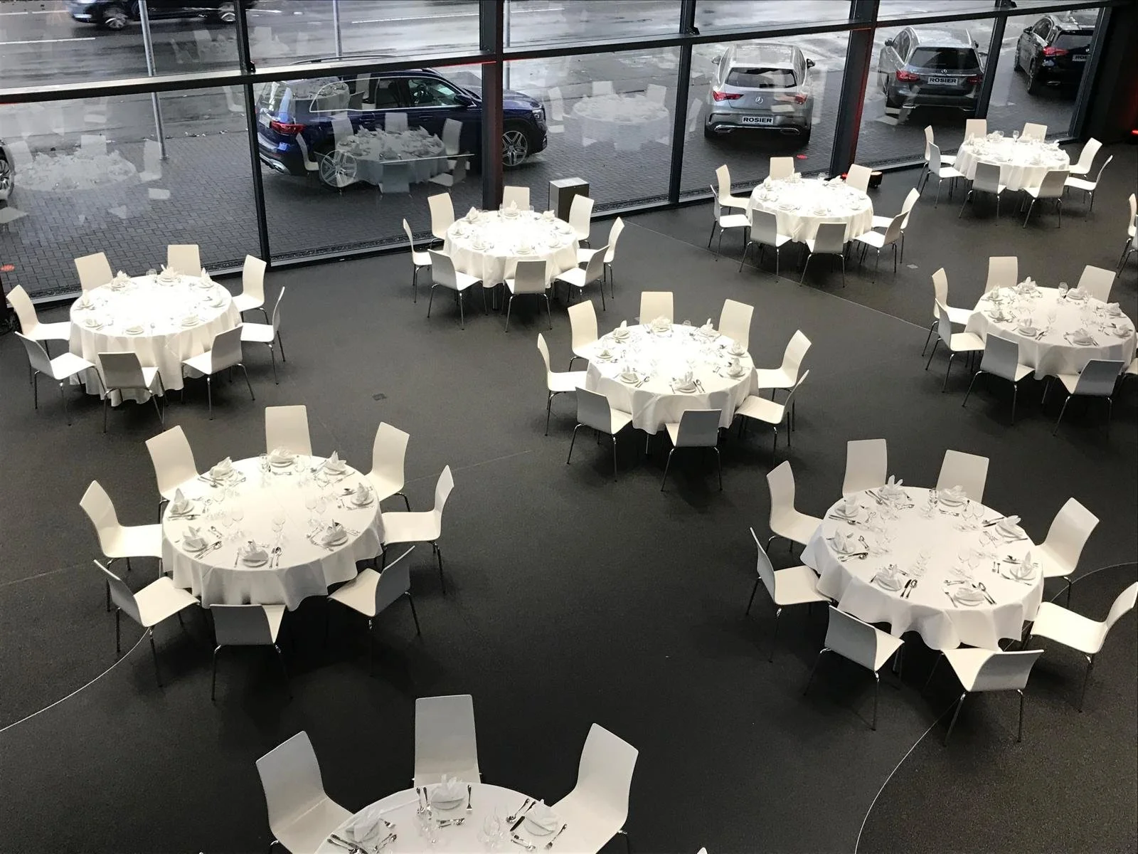 Empty banquet hall with round tables covered in white tablecloths, set with utensils, plates, and glasses, surrounded by white chairs, with large windows showing parked cars outside.
