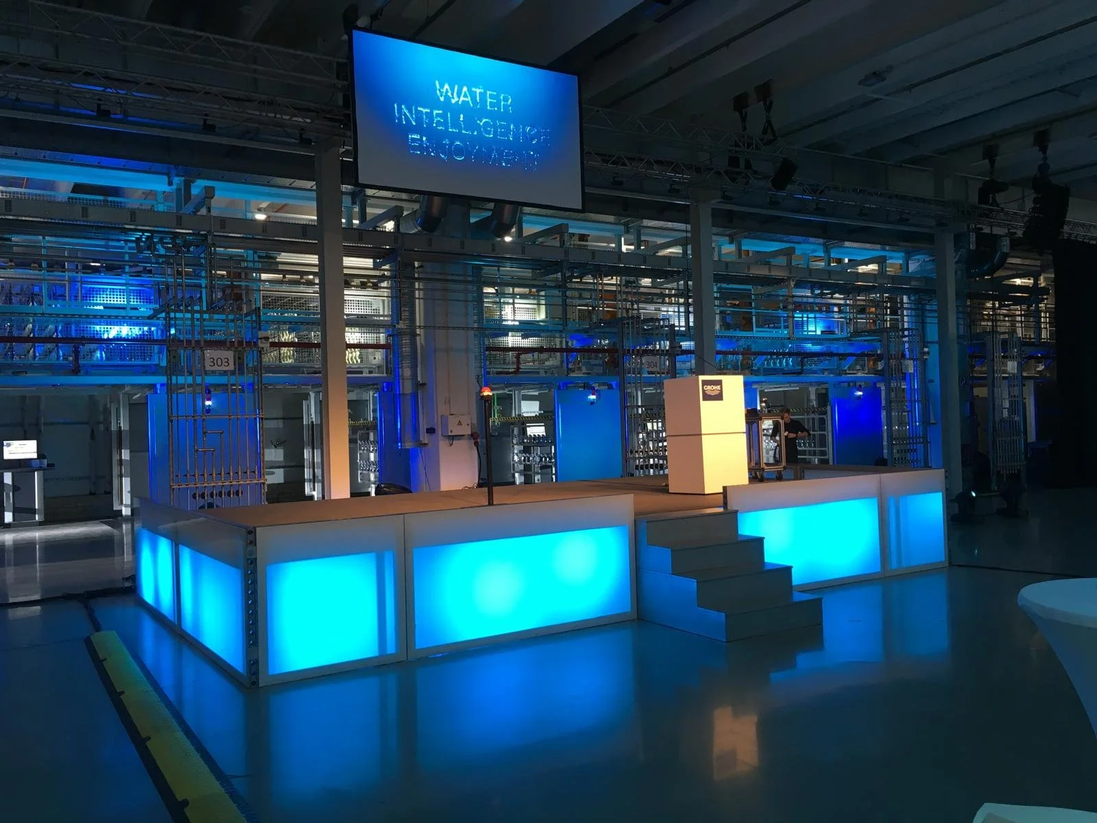 An indoor exhibition space with a stage, has a sign reading 'Water, Intelligence, Environment,' illuminated in blue. Behind the stage is a complex metal framework with pipes and machinery, and a person is seen near the right side of the stage.