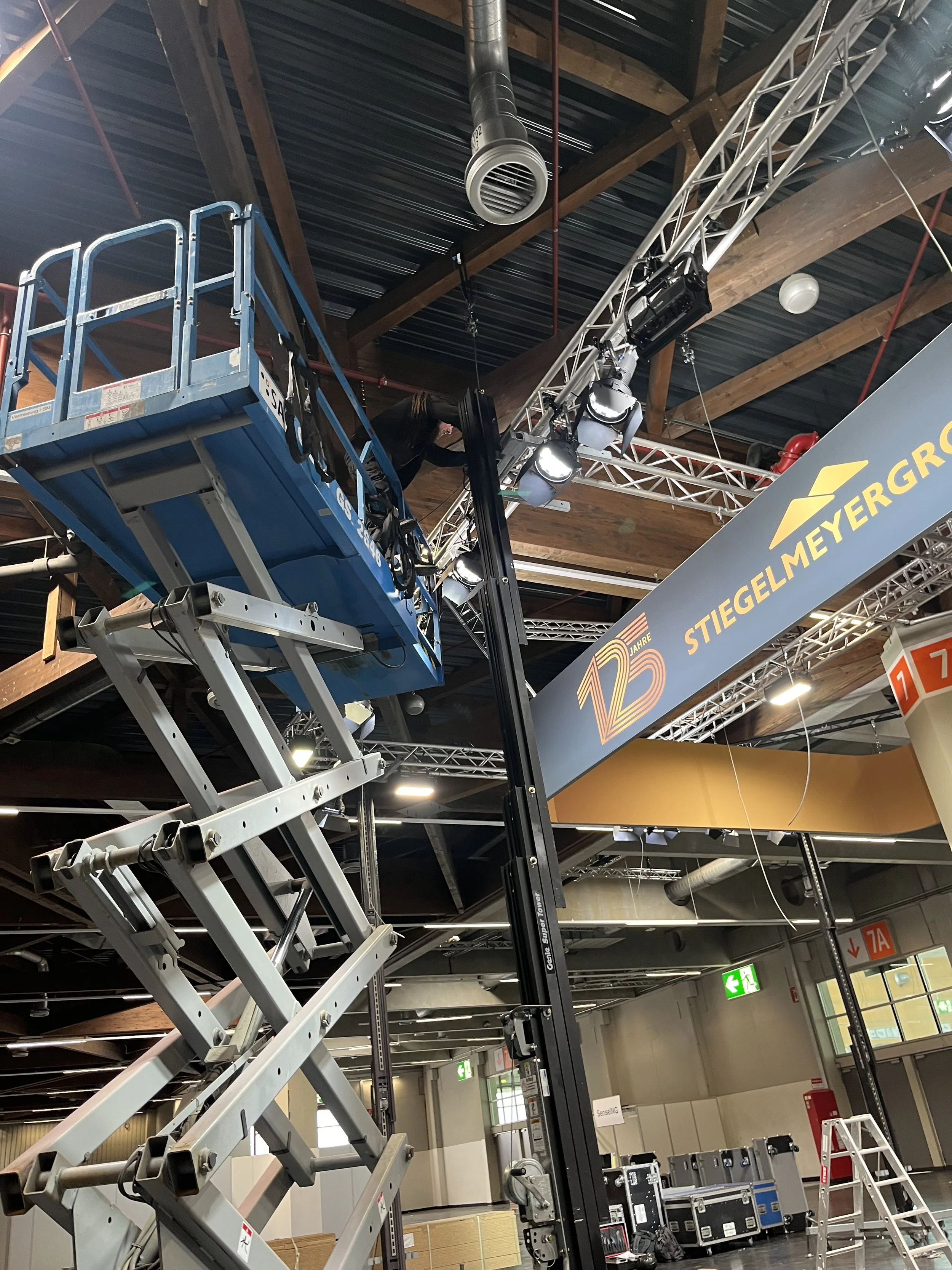 Indoor event setup with a scissor lift, truss system, and stage banner celebrating 125 years of Stieglem MeyerGrol. Equipment and ladders are visible in a large open space.