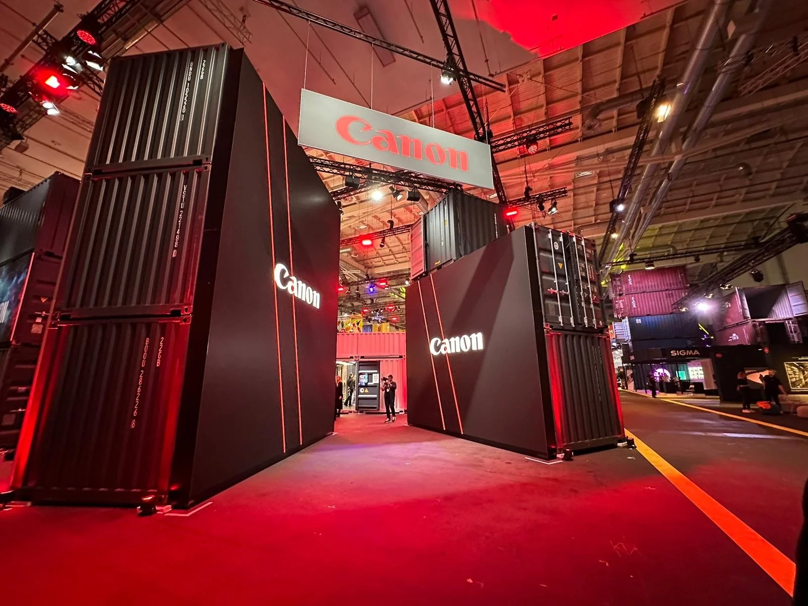 Exhibition booth showcasing Canon products with large black shipping containers and prominent red and white Canon signage, illuminated by red and white lights, inside a spacious trade show hall.