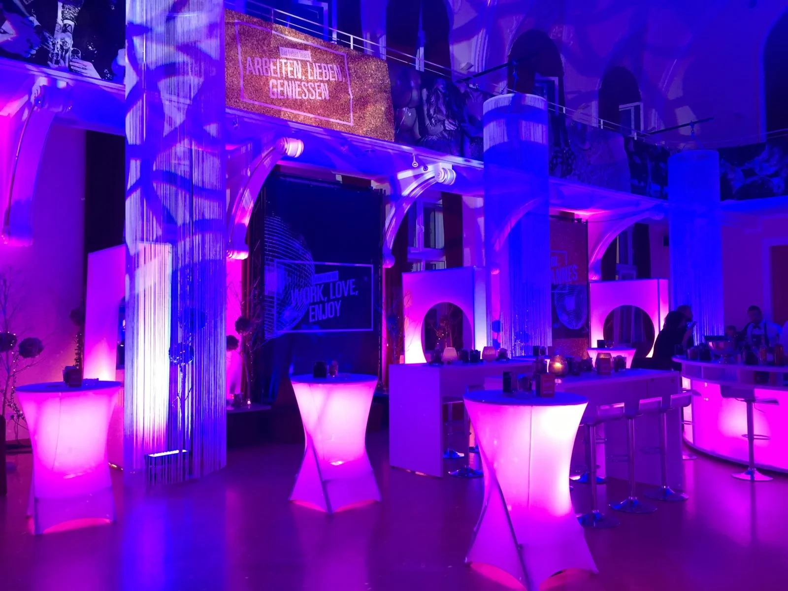 A dimly lit event space with purple and pink lighting, high tables, and bar counters, decorated with signs and modern decor, in an elegant venue.
