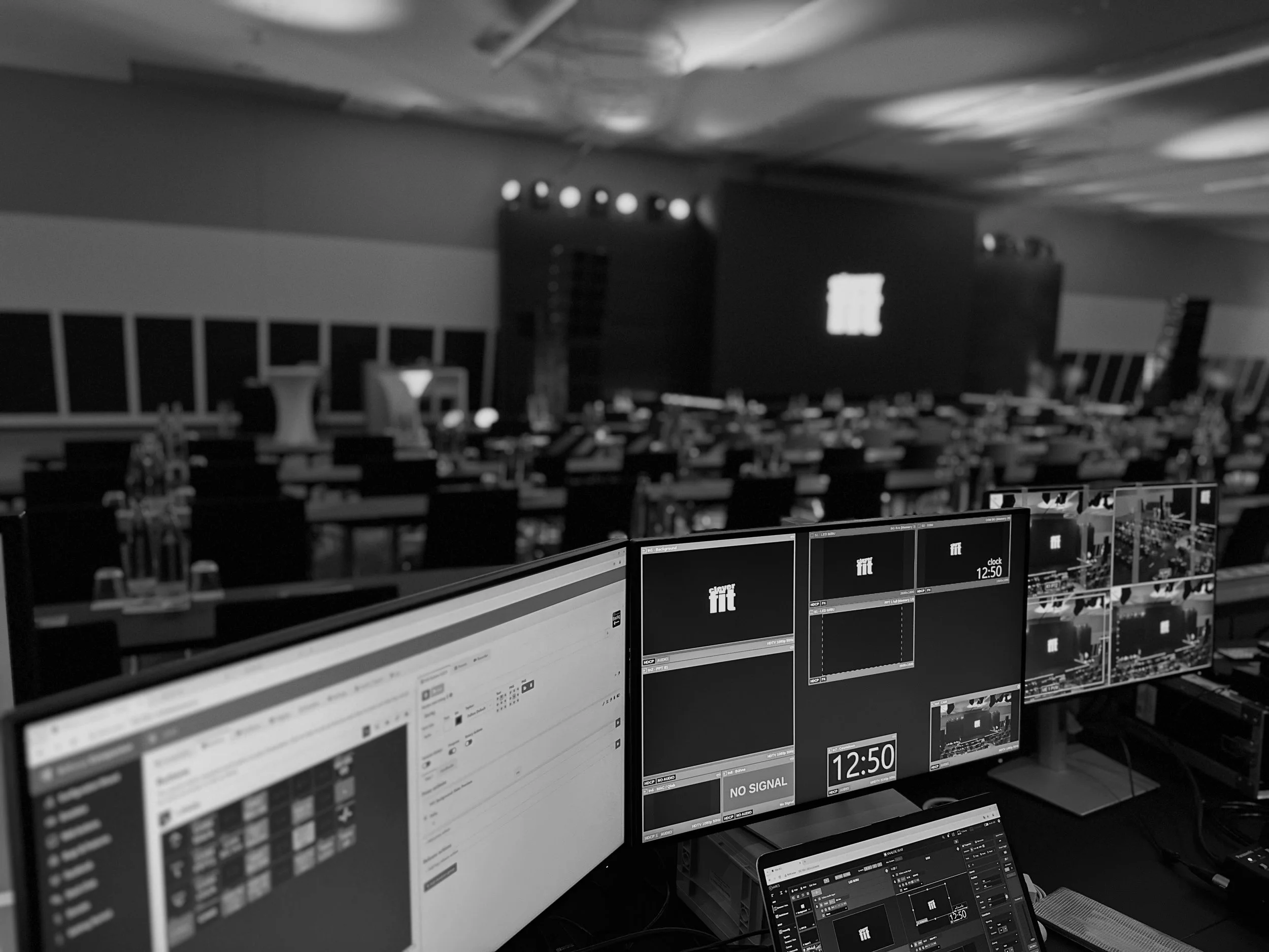 A control room with multiple monitors displaying schedules, clocks, and broadcast information in front of empty chairs and tables, with a stage and large screen in the background.