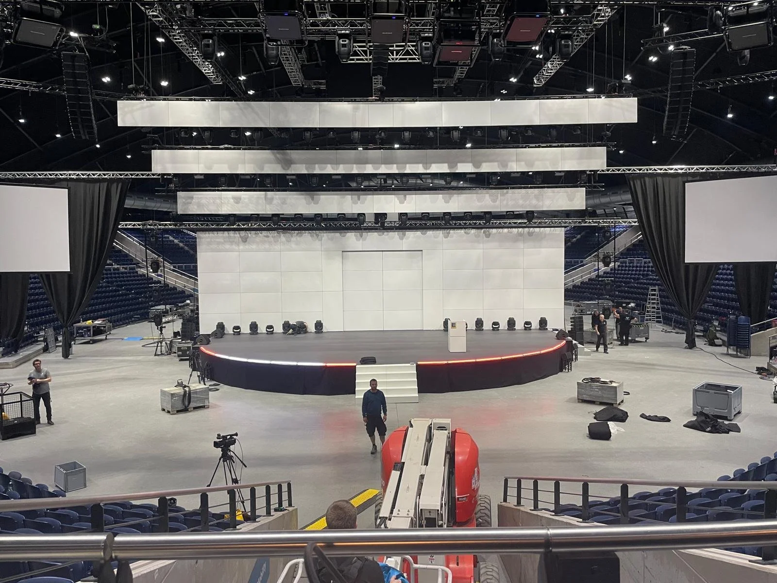 An empty stage with a large backdrop, surrounded by lighting equipment, large screens, and crew members setting up in an arena. There is a curved platform in front of the stage, and various cables and boxes scattered around.