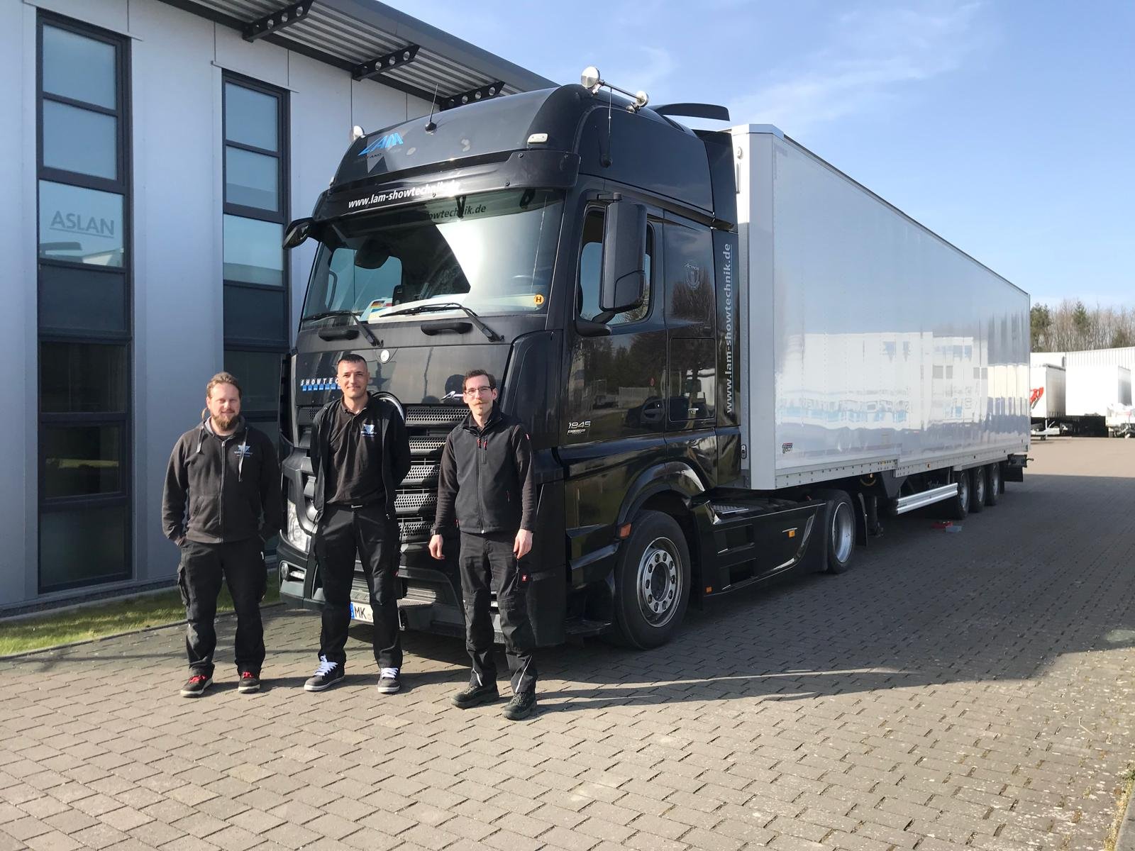 Three men standing beside a large black and white truck outside a building with glass windows.