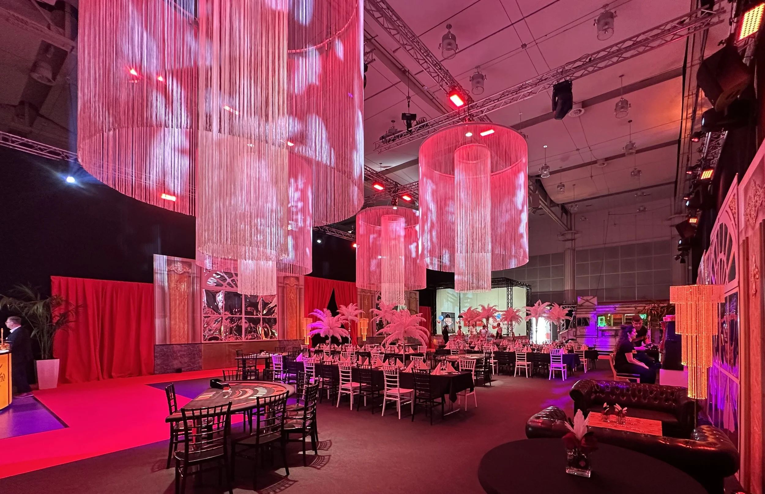 Interior of a decorated event venue with pink lighting, large hanging pink chandelier lights, pink palm trees, and various seating arrangements including black and white chairs and a black couch.