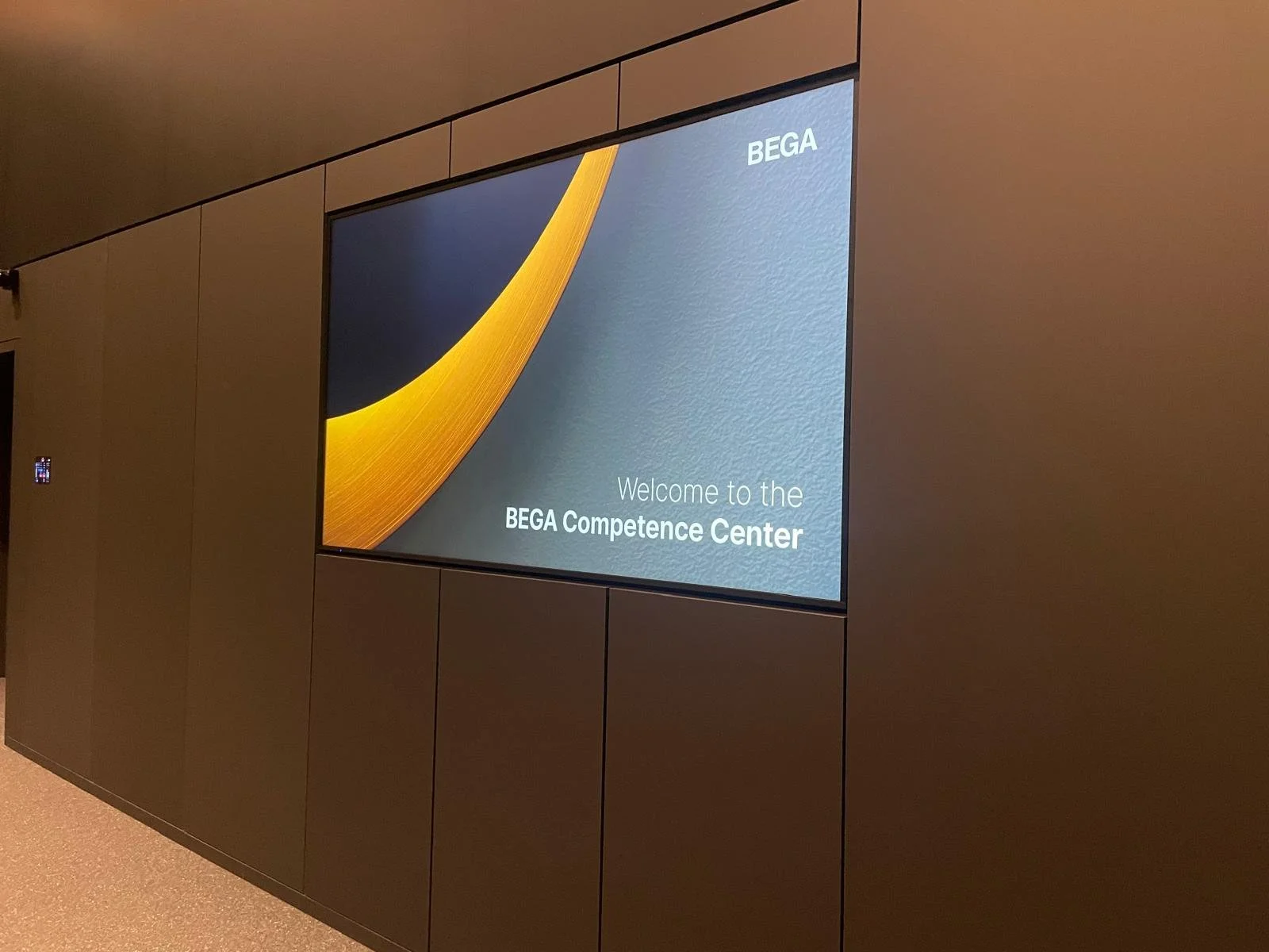 Digital display on a wall, welcoming visitors to the BEGA Competence Center with a blue background, a yellow curved graphic, and white text.