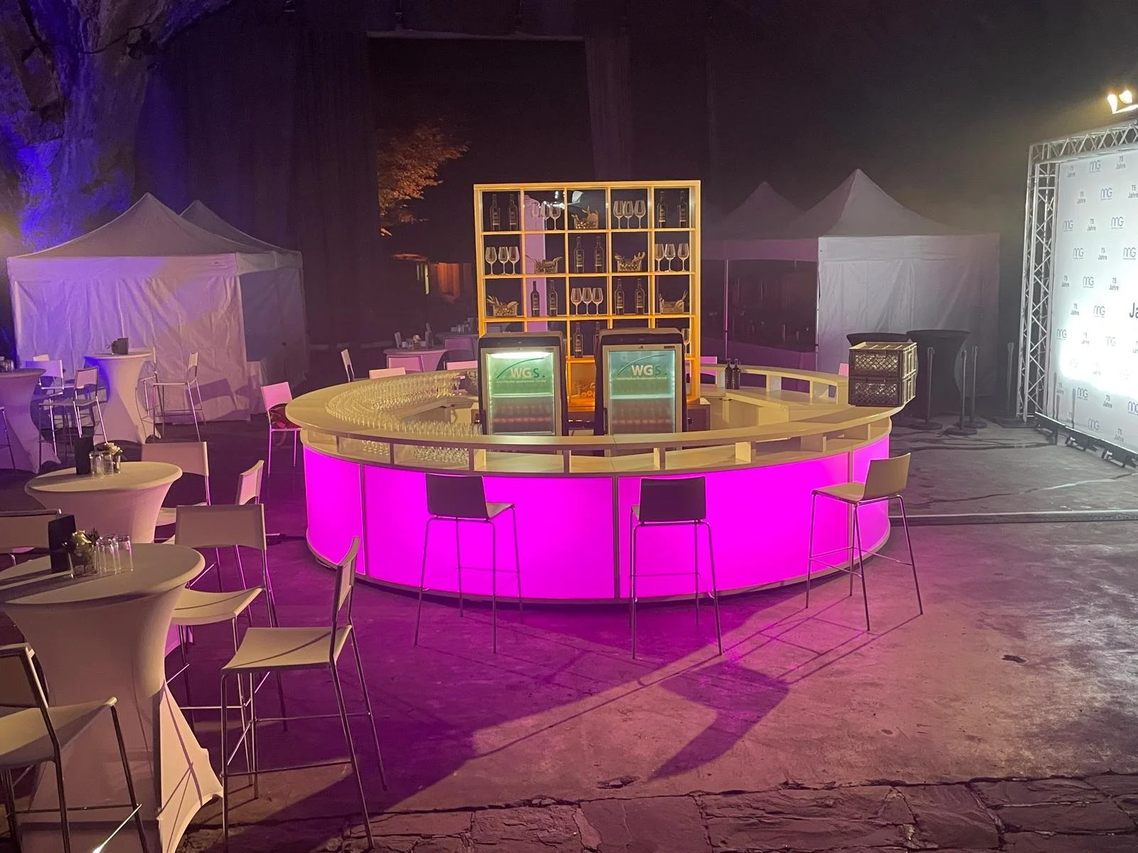 A nighttime outdoor event setup featuring a brightly lit pink circular bar with two drinks dispensers on top, surrounded by high chairs. Behind the bar is a yellow shelving unit display with glasses and bottles, with white tents and tables with chair