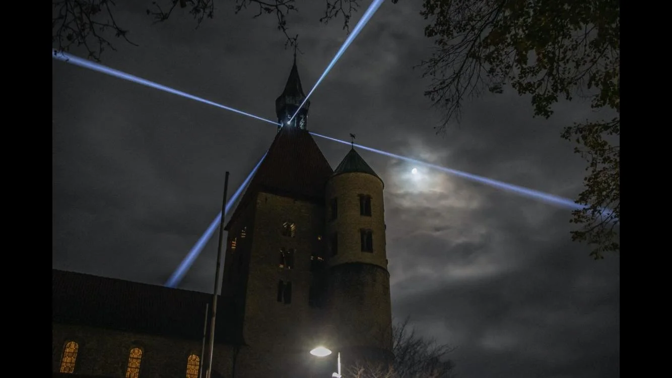 Nighttime view of a church with light beams emanating from its steeple, the moon partially obscured by clouds, and trees framing the scene.