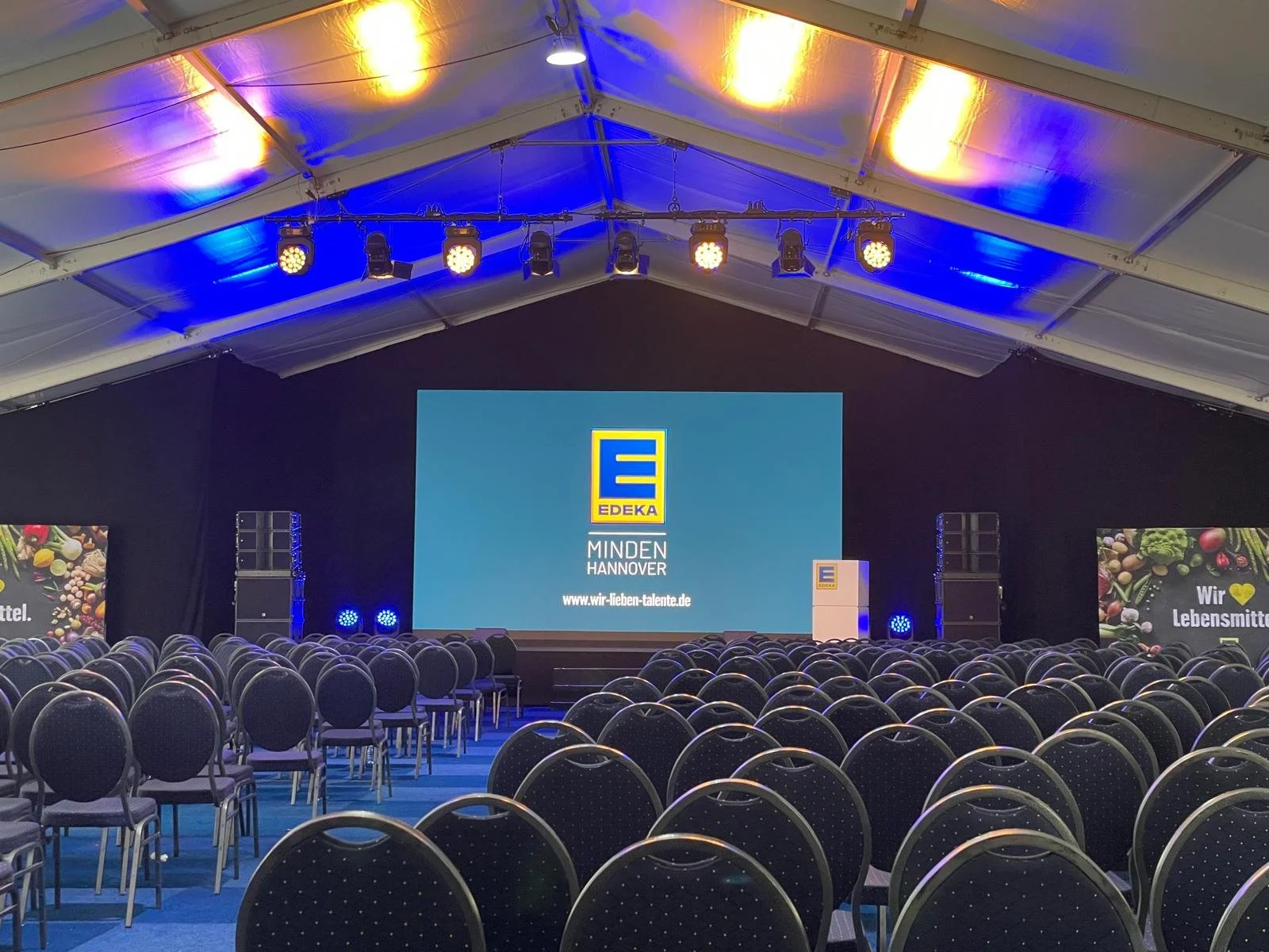 Empty conference room with rows of chairs facing a stage with a large screen displaying the EDEKA logo, with colored lighting and side screens showing food images and text.