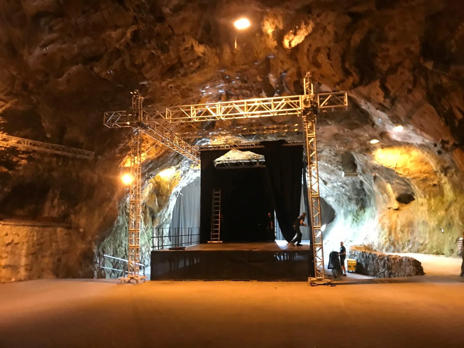 An indoor stage set up inside a large rocky cavern with a black curtain, scaffolding, and lighting gear.