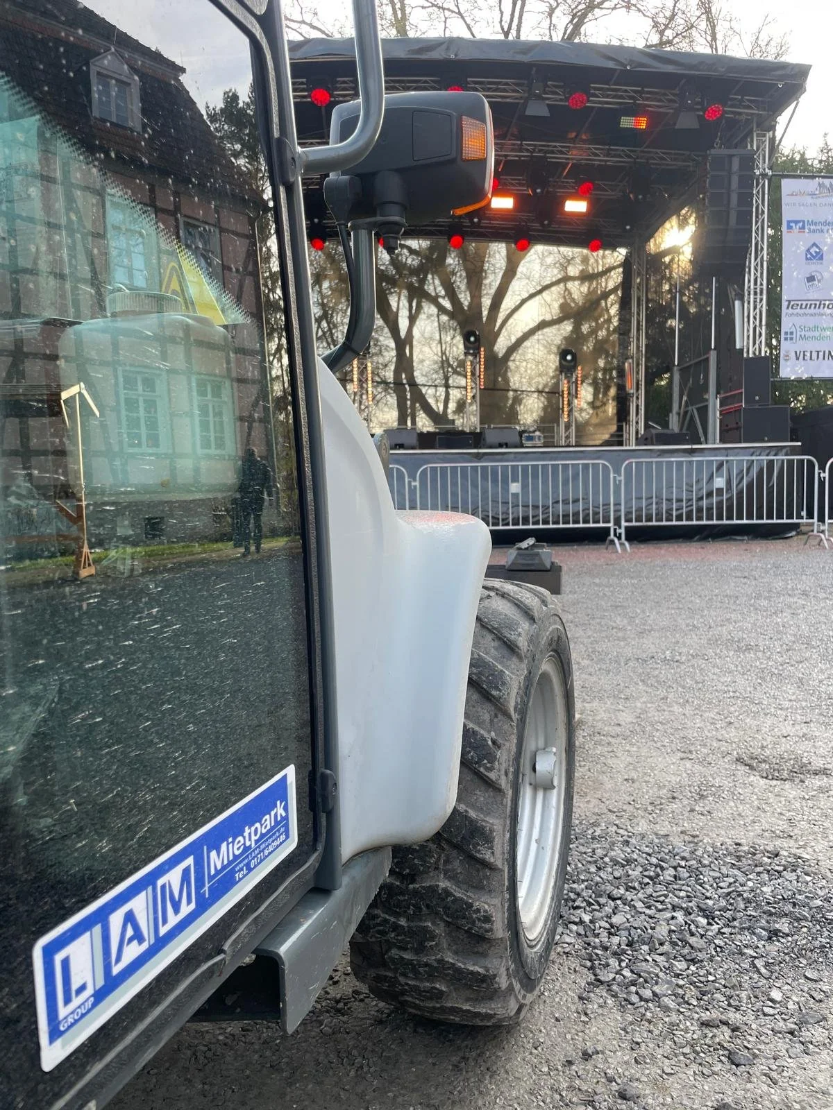 Close-up of a construction vehicle parked in front of an outdoor stage with sound and lighting equipment, trees, and a sunset in the background.