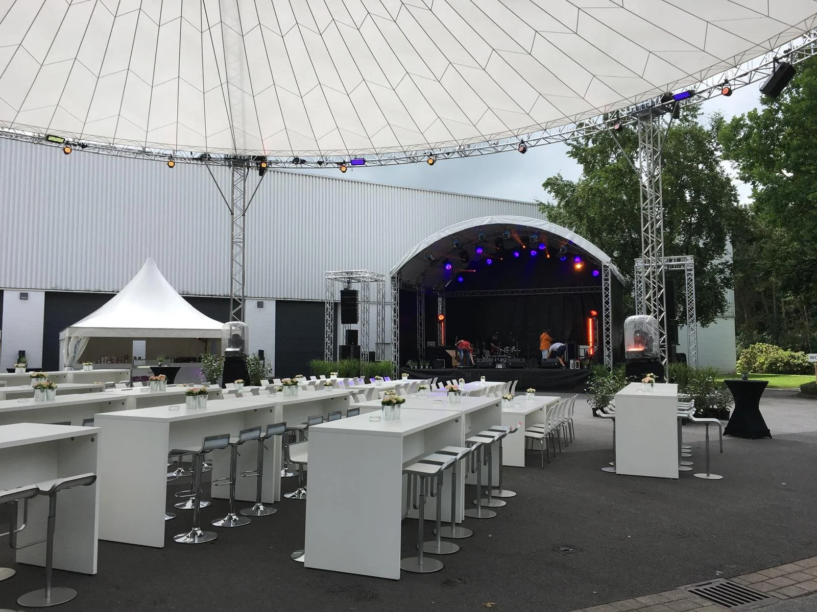 An outdoor stage set up with lighting and sound equipment, under a large canopy. White high tables with flower arrangements and barstools are arranged in front of the stage, with a small white tent nearby. The scene suggests preparations for a live o
