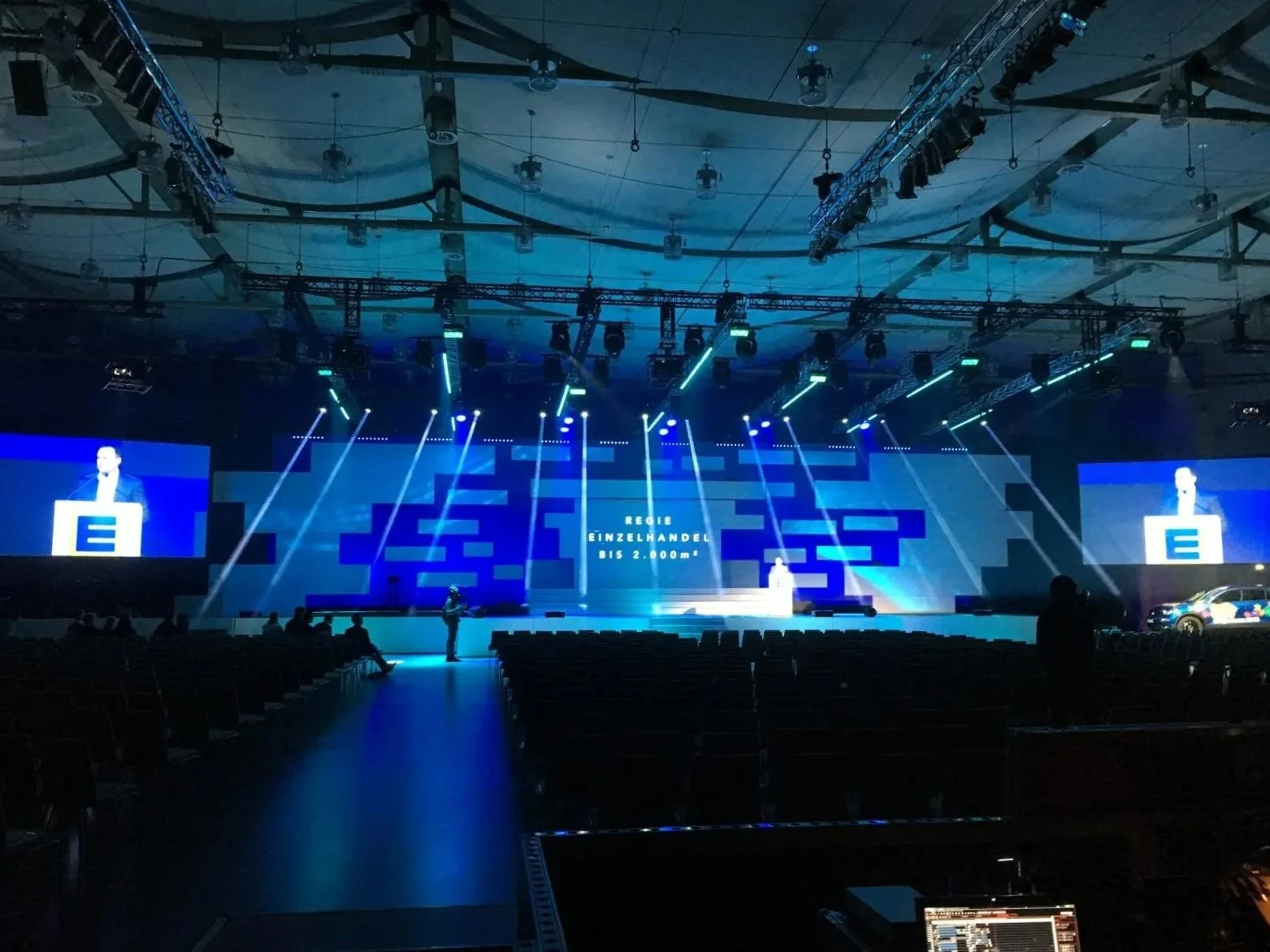 An empty conference hall with a stage featuring large screens and blue lighting, preparing for an event or presentation.