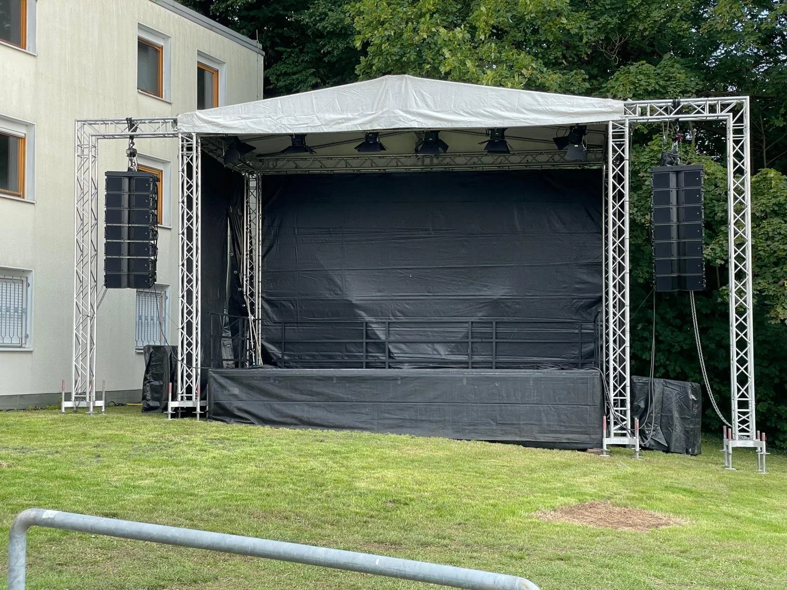 An empty outdoor stage with a black backdrop, metal support frame, and two large black speaker systems hanging on each side, set on a grassy area with trees and a building in the background.