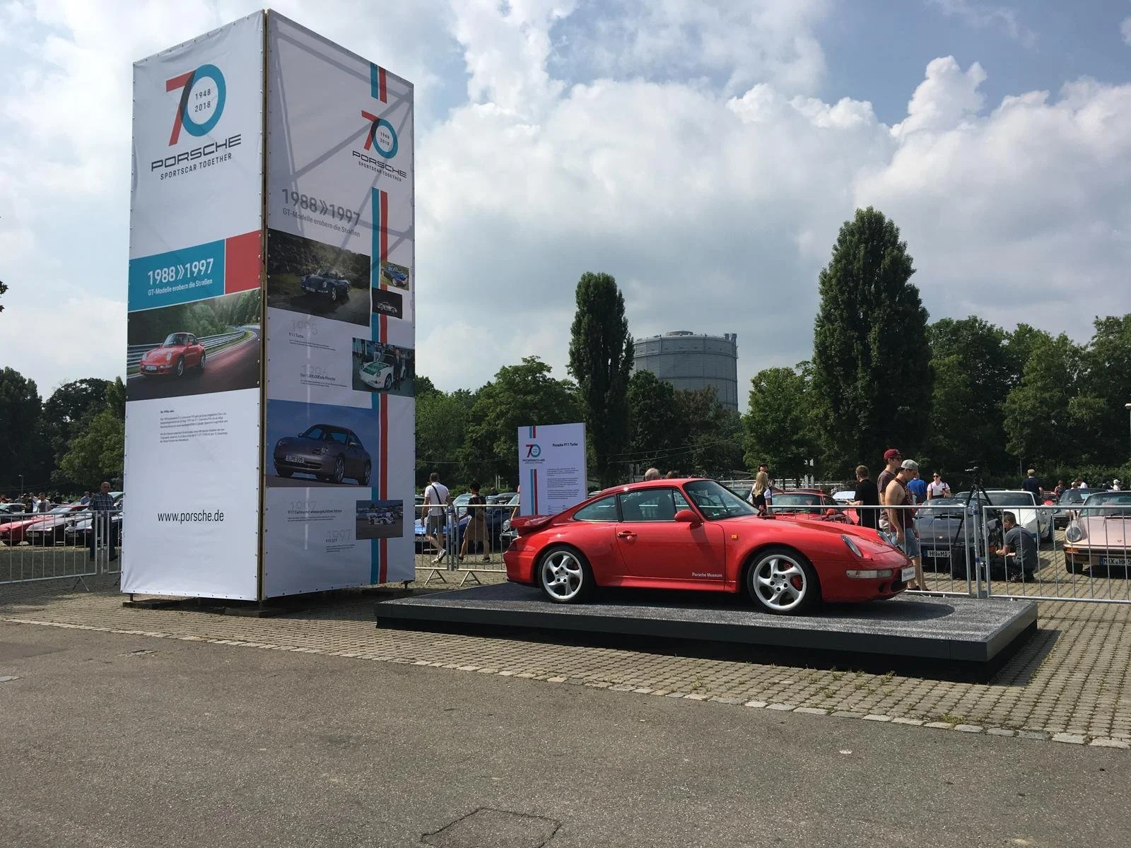 Red Porsche sports car displayed on a platform at an outdoor car exhibition, with informational banners and a crowd of people in the background.