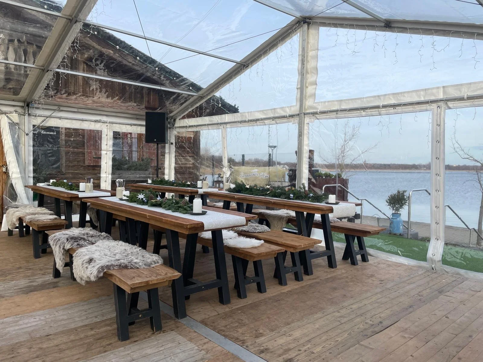 Festively decorated outdoor dining area with wooden tables and benches, fur throws, Christmas garland, candles, and a view of water outside a transparent tent.