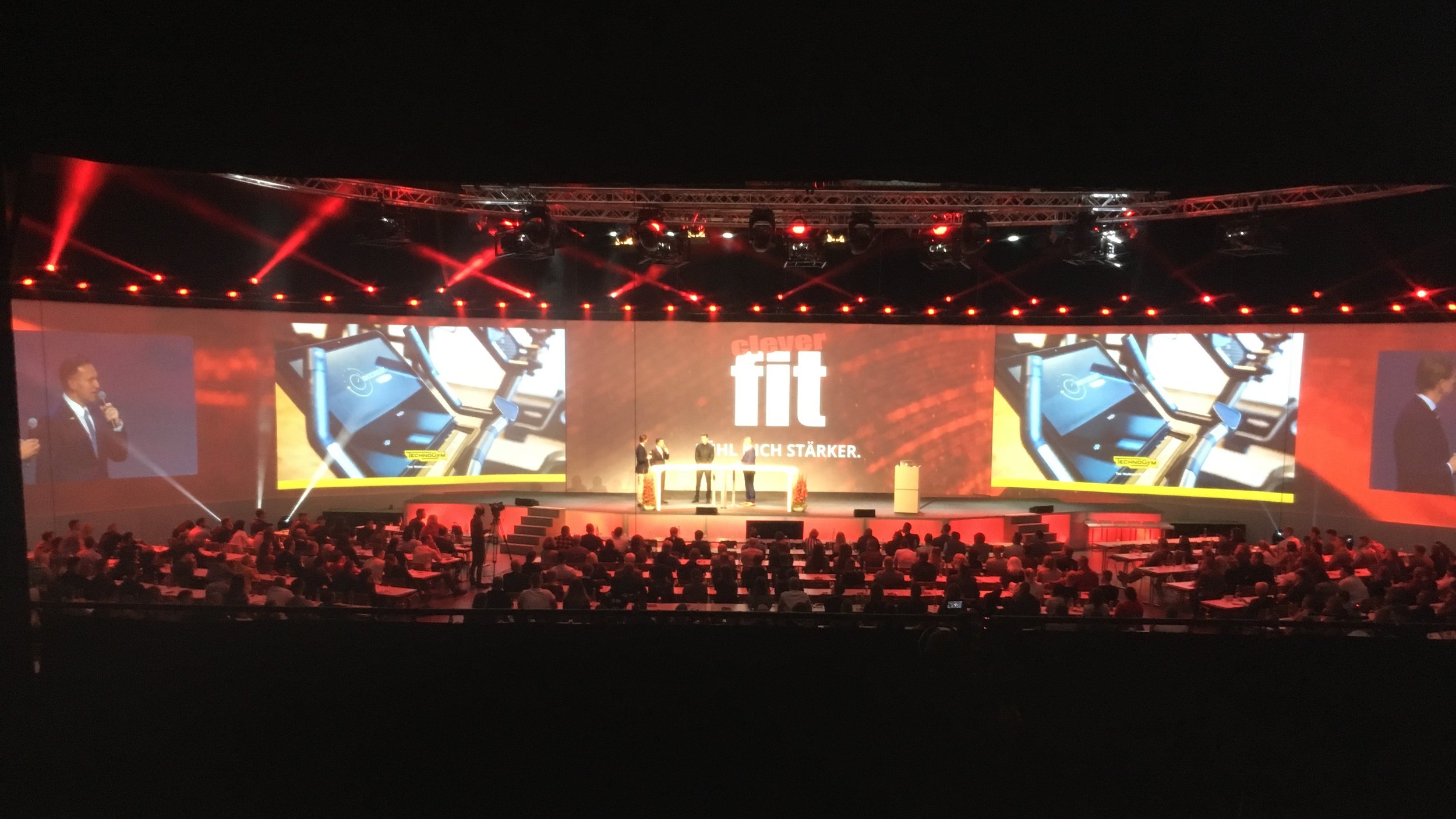 A large conference or event stage with a backdrop displaying the brand 'fischer fit'. The stage features multiple screens showing close-up images and a presenter. The audience is seated facing the stage, and there are red stage lights overhead.