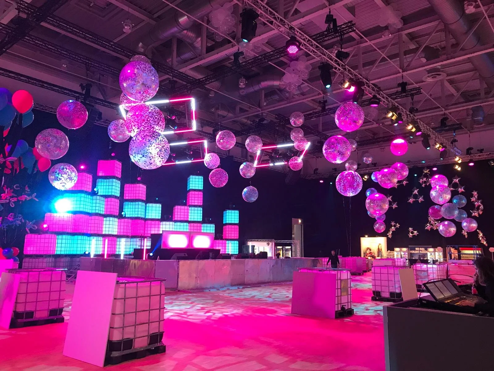 An indoor event space decorated with pink, purple, and blue lights, featuring hanging spherical and star-shaped balloons, illuminated cube structures, and a dance floor area.