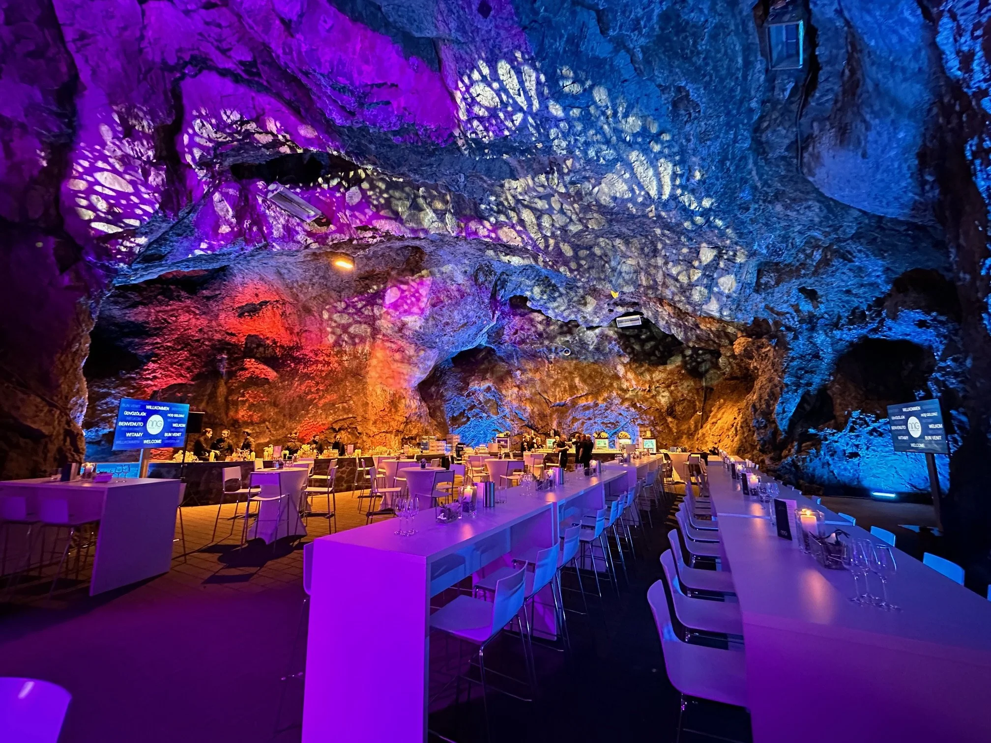 An underground cave transformed into a restaurant or event space with tables set for dining, colorful lighting illuminating the rocky walls and ceiling.