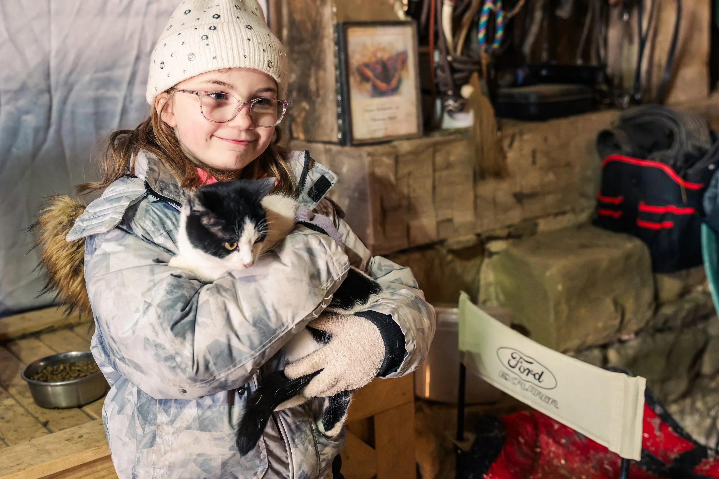 A young girl wearing glasses, a beige knit hat, and a winter coat is holding a black and white cat in a cozy indoor setting. She is smiling gently and looking at the camera.
