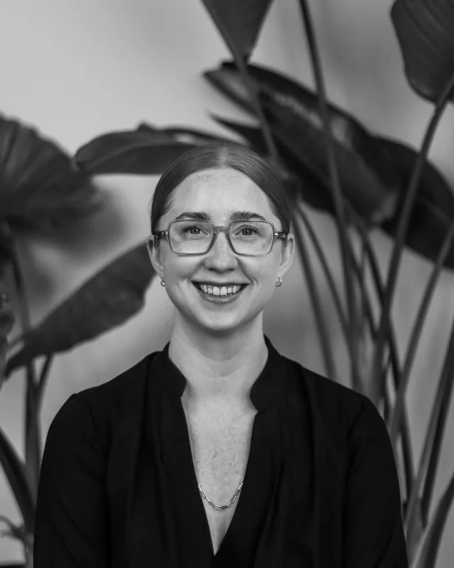 A woman with glasses smiling in front of large leafy indoor plants.